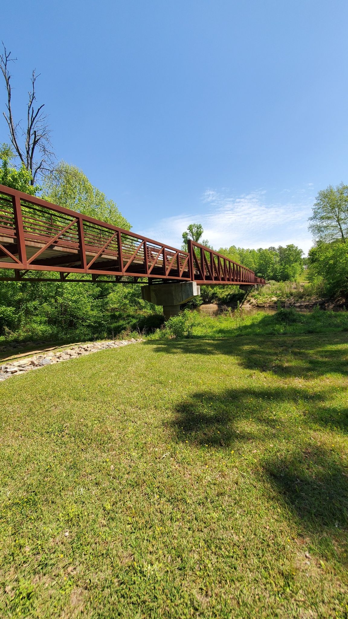 A long, rusty metal bridge spans over green grass under a clear blue sky. - Neuse River Pedestrian Bridge – Neuse River Greenway loop from Perry Creek Rd at Capital Blvd