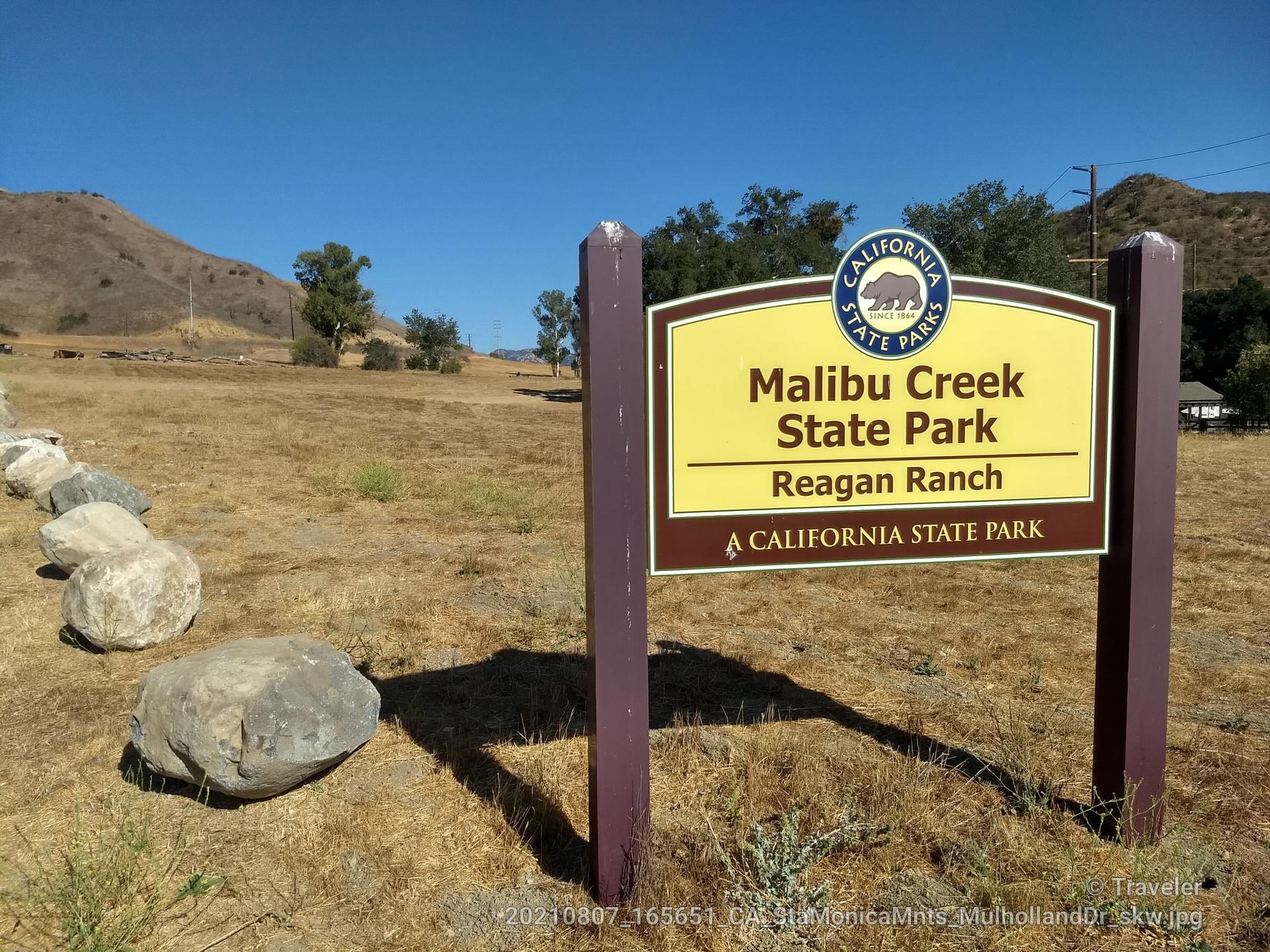 Wooden park sign and large rocks in a sunny, dry, hilly landscape.