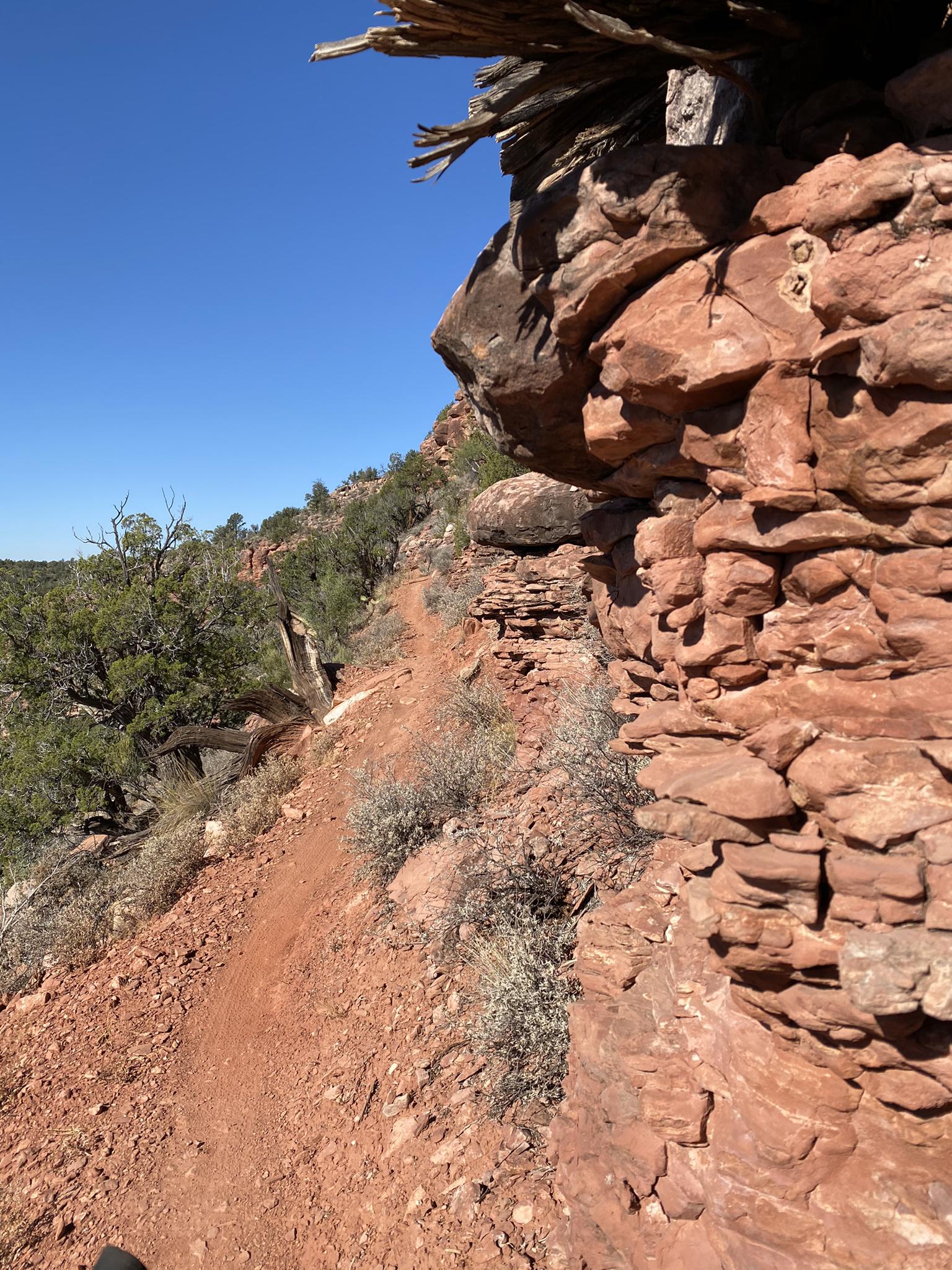 Narrow dirt trail winding alongside a towering red rock cliff.