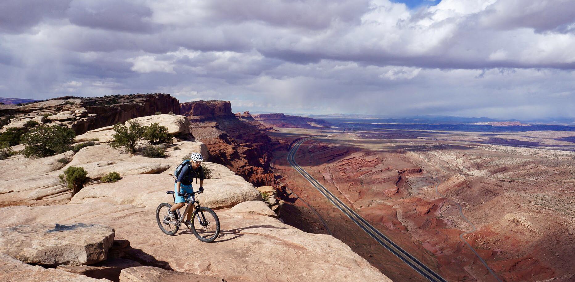 Mountain biker stands on a cliff overlooking a vast red rock canyon with a road below.