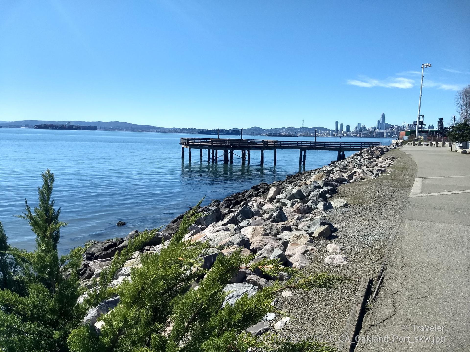 Calm bay with a long pier, rocky shore, and distant city skyline under blue sky.
