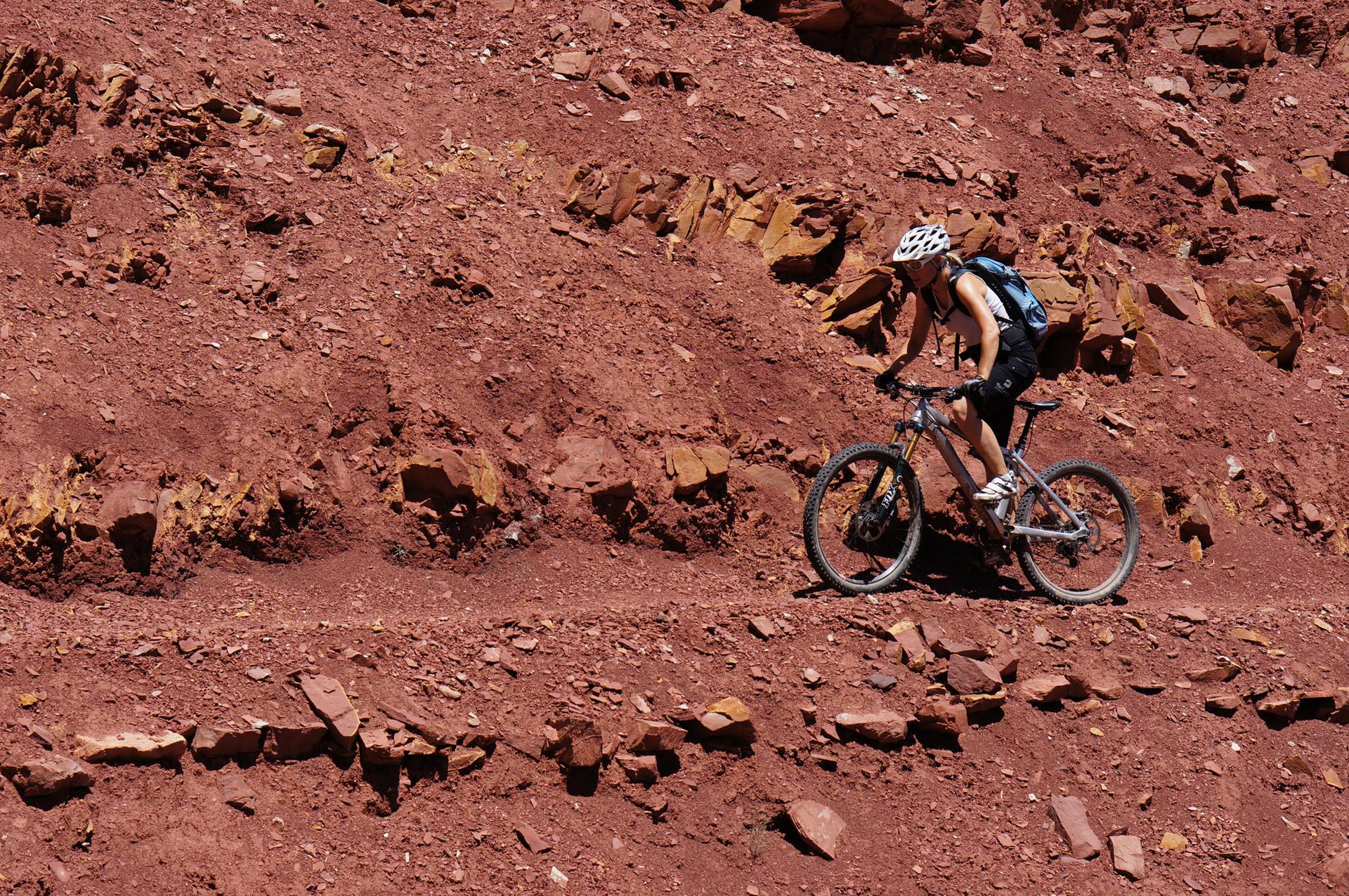 Person mountain biking on a red rocky trail under bright daylight.