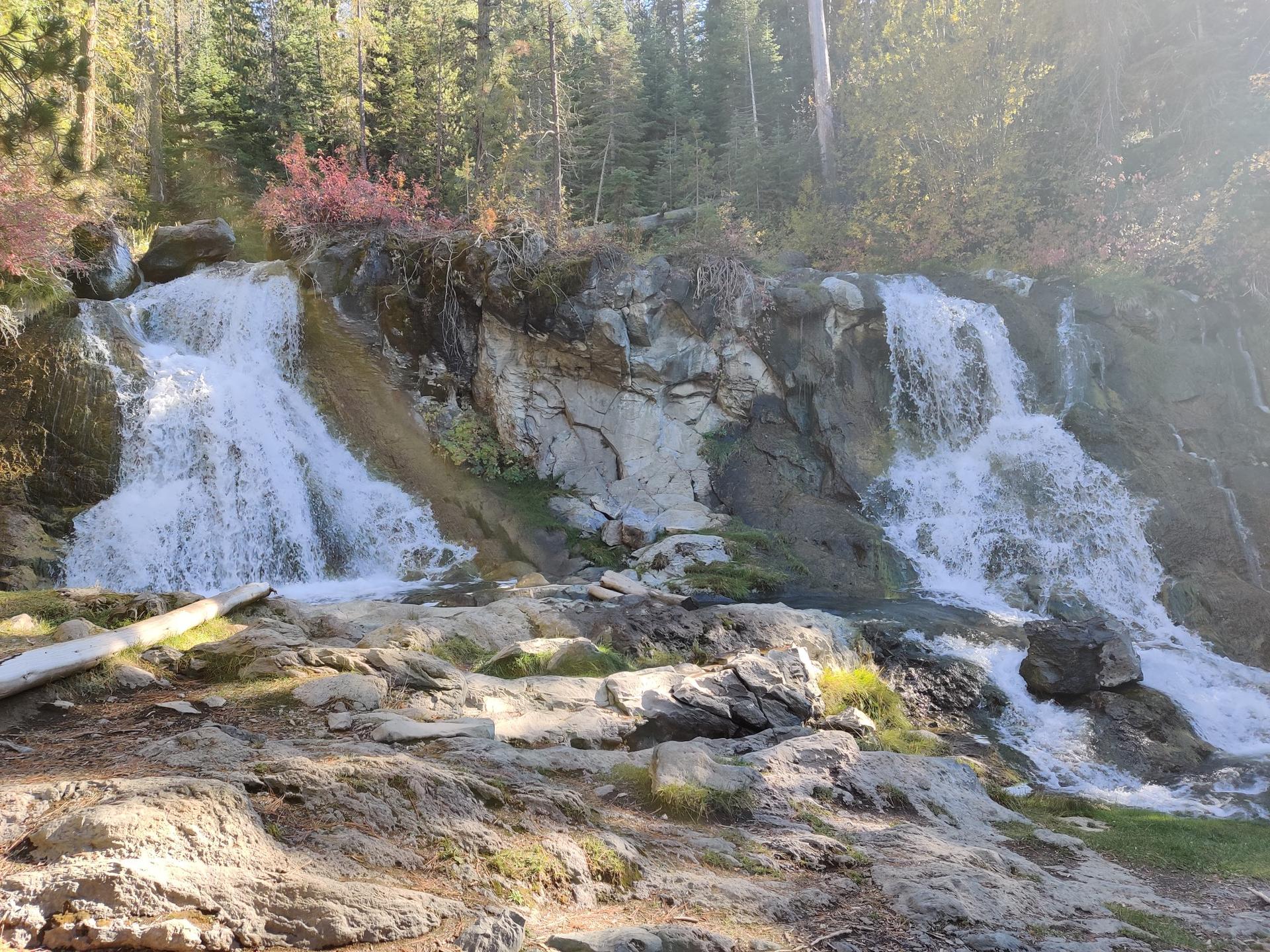 Two waterfalls cascade over mossy rocks with trees in bright sunlight.