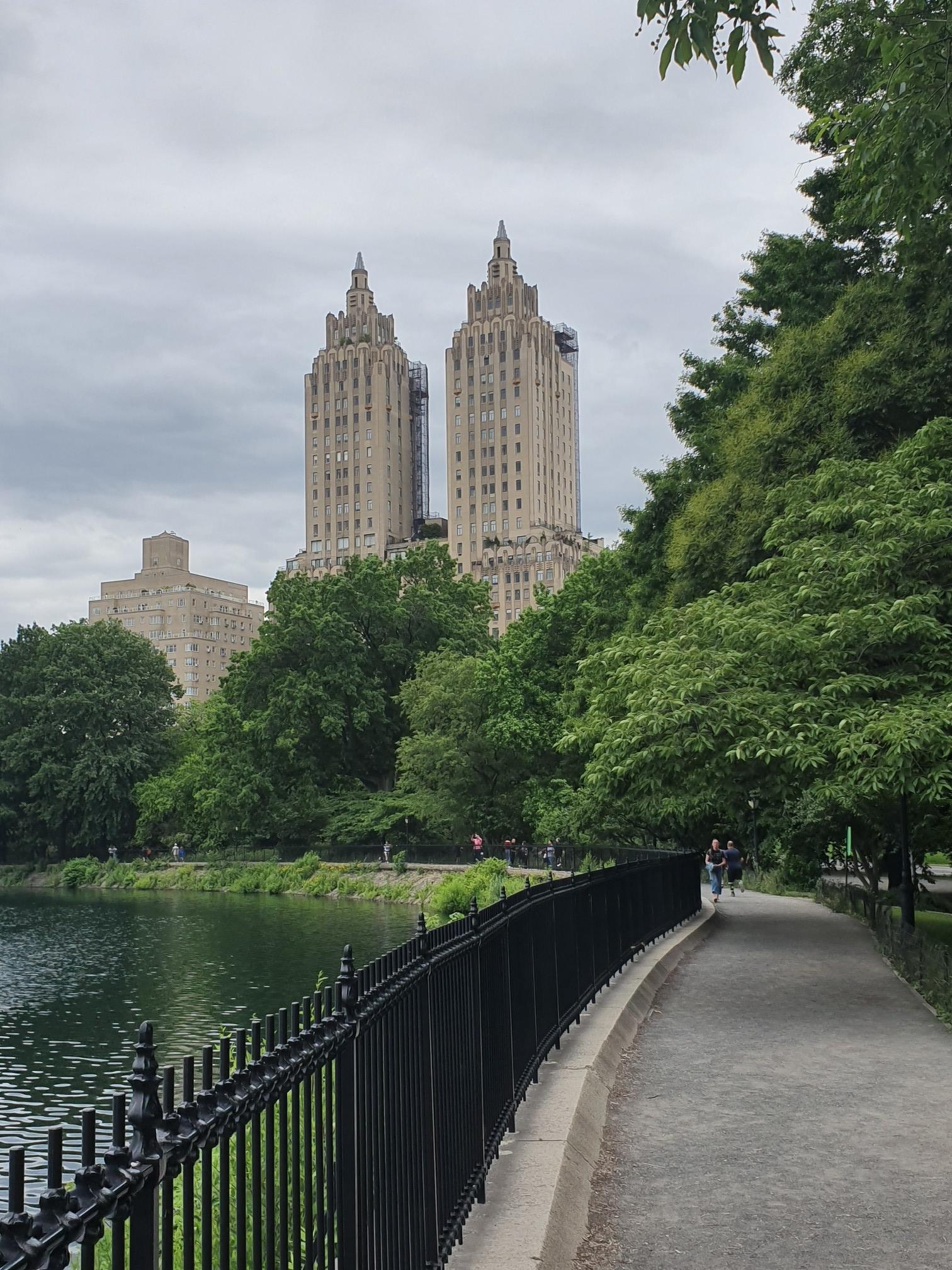 Path next to a lake with tall buildings and trees under an overcast sky.