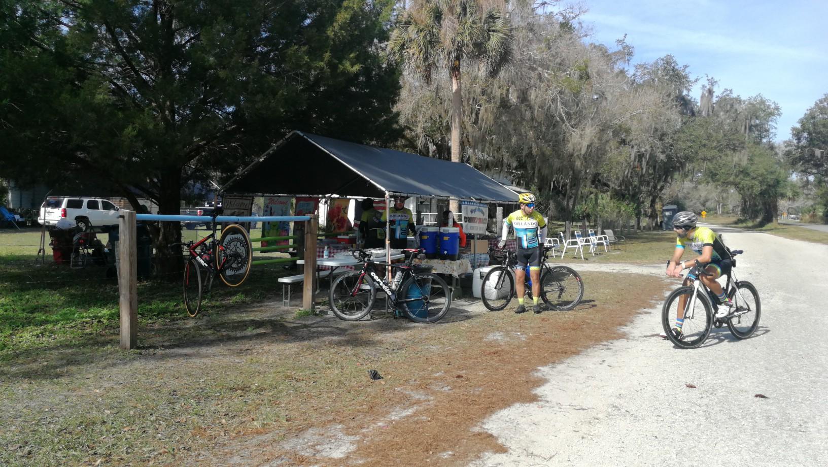 Cyclists gathered under a canopy next to a dirt path on a sunny day.