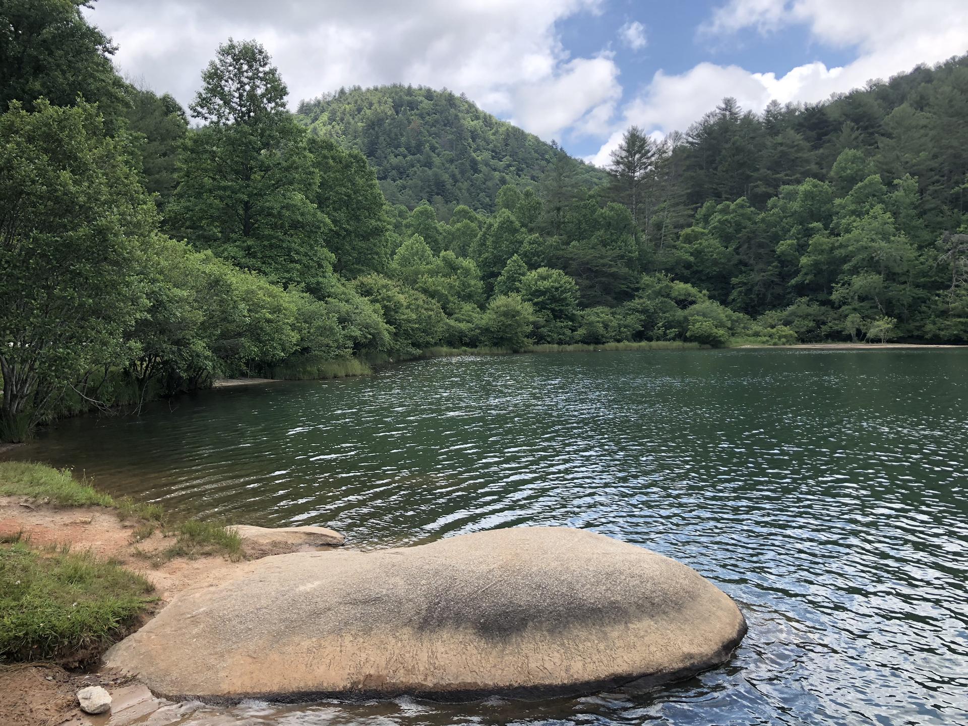 Lake with a large smooth rock in the foreground, surrounded by forested hills.