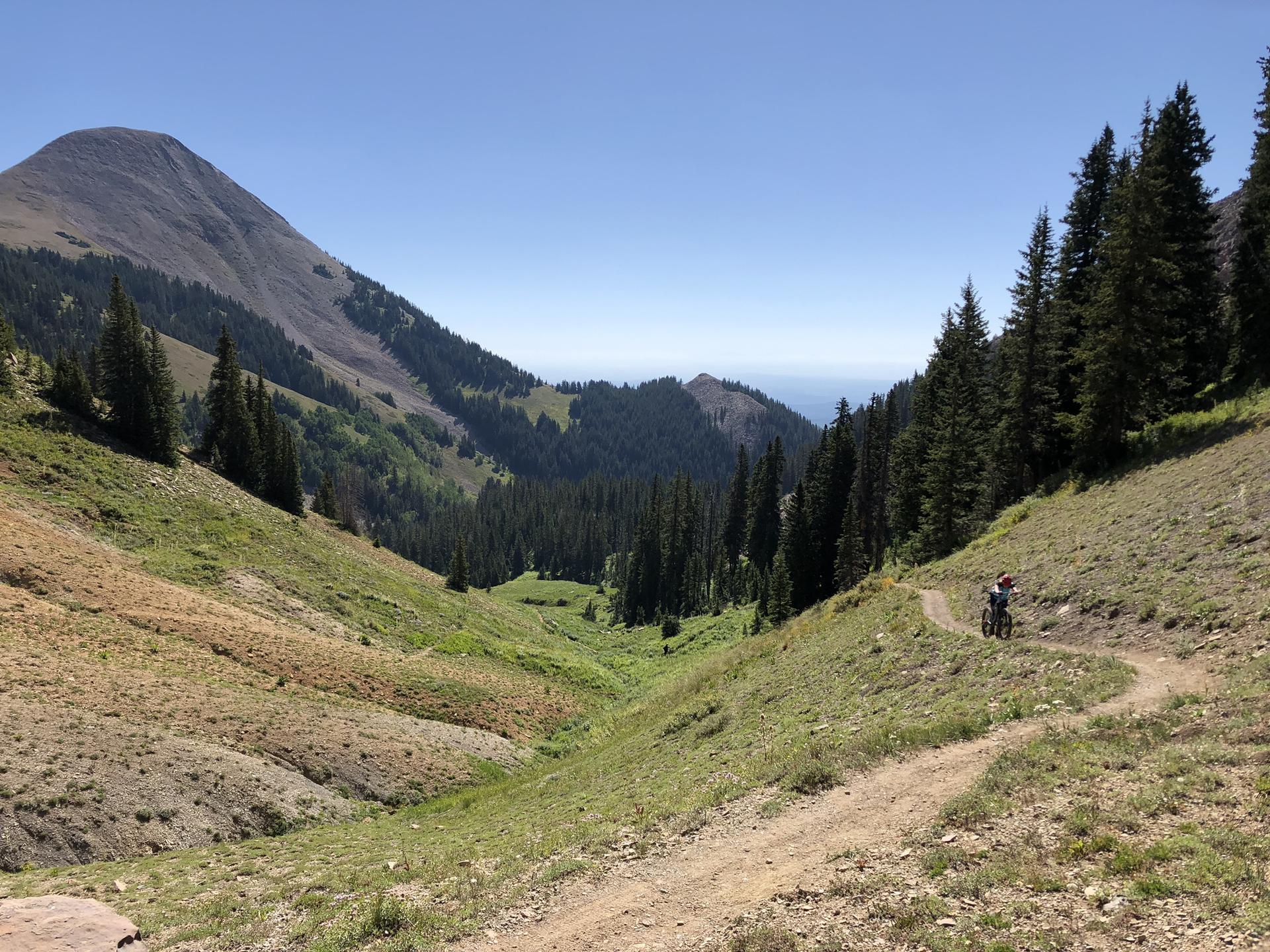 Mountain biker on a trail in a green mountain valley under clear skies.