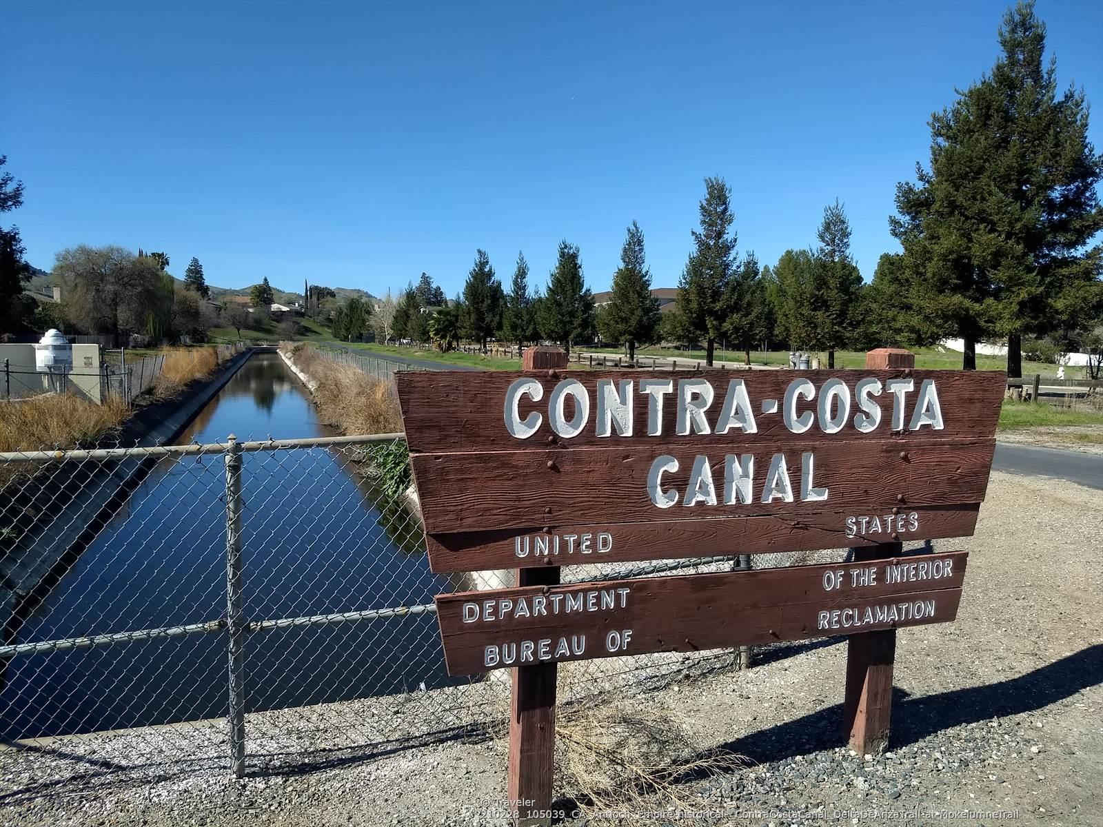 A large wooden sign stands beside a canal and fence on a sunny day.