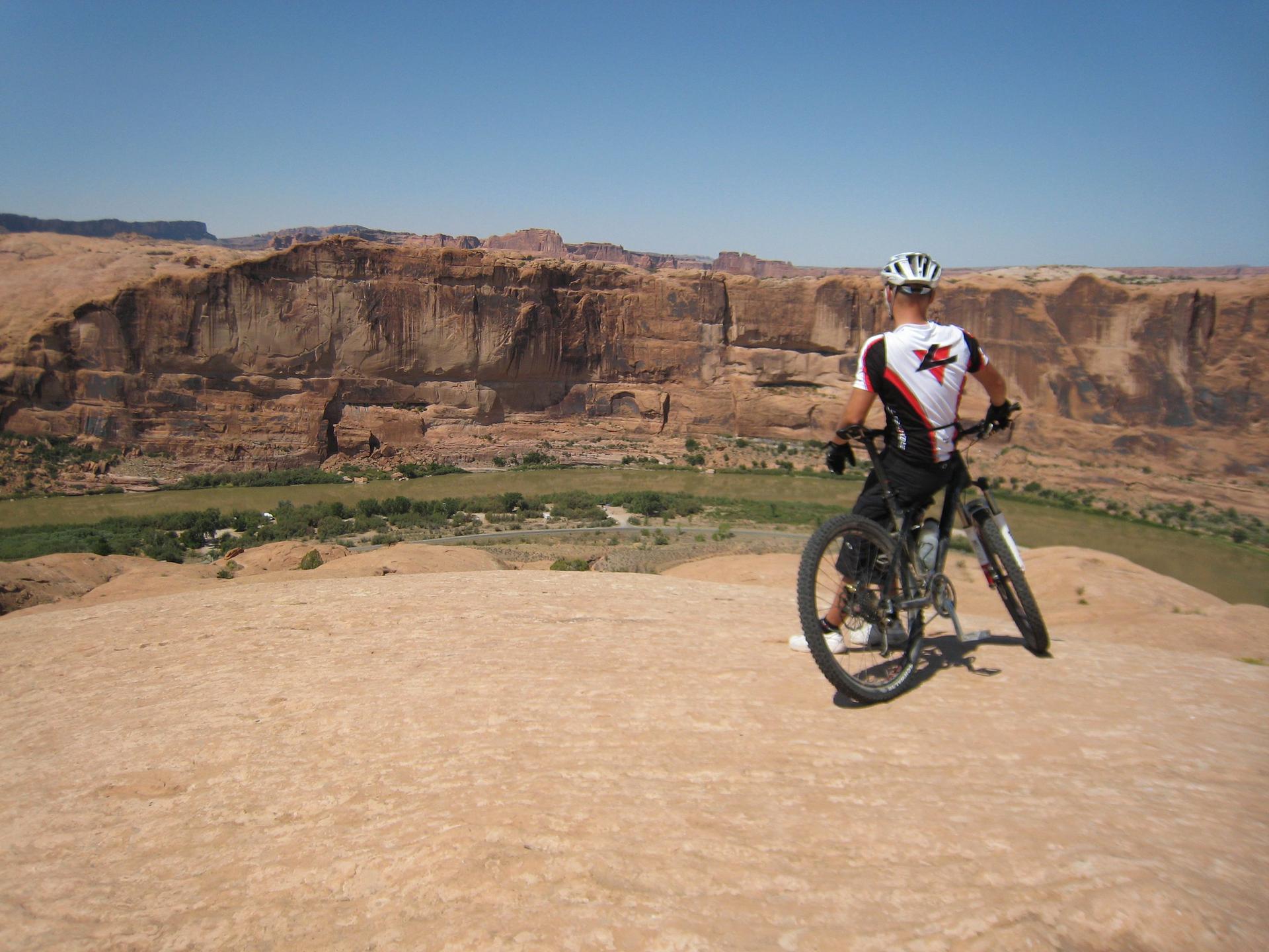 Cyclist on a rocky cliff overlooking a green valley and red formations.