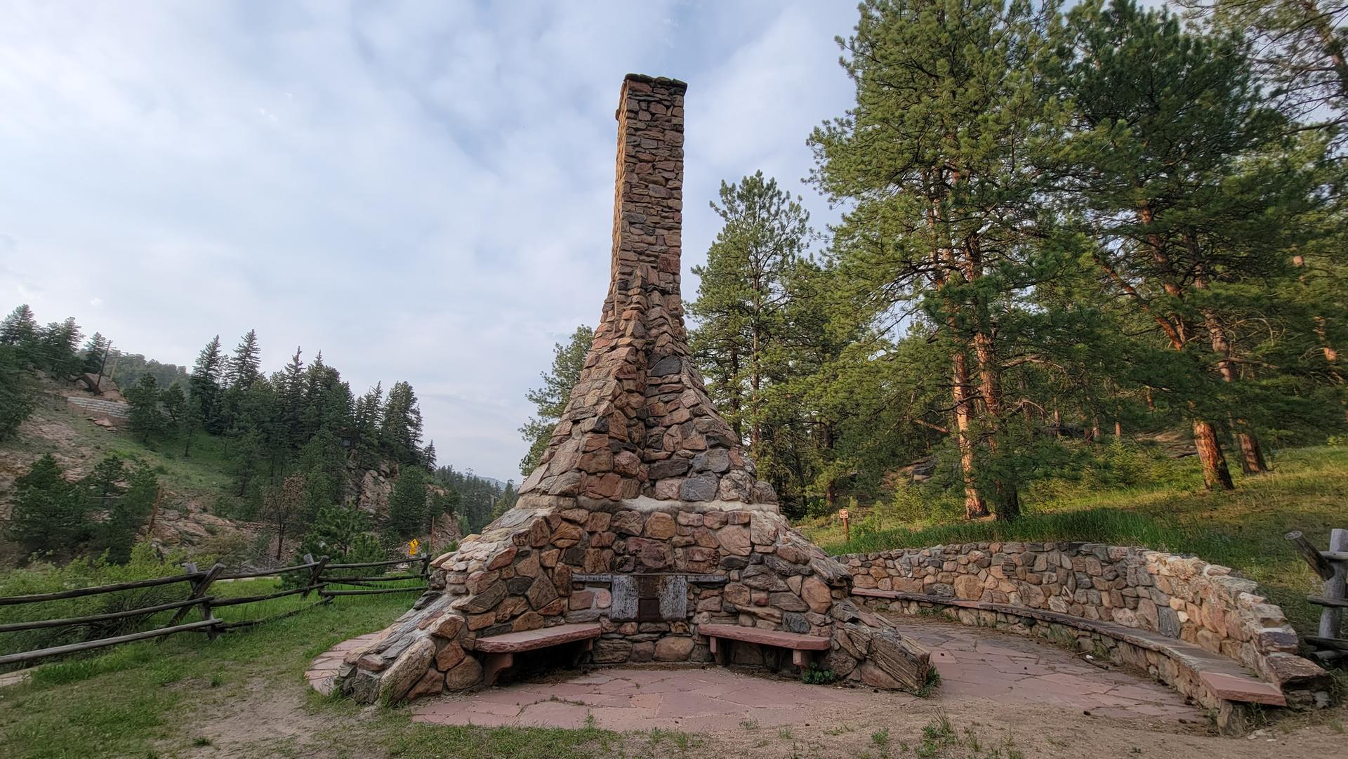 A tall stone chimney structure with benches, surrounded by pine trees.