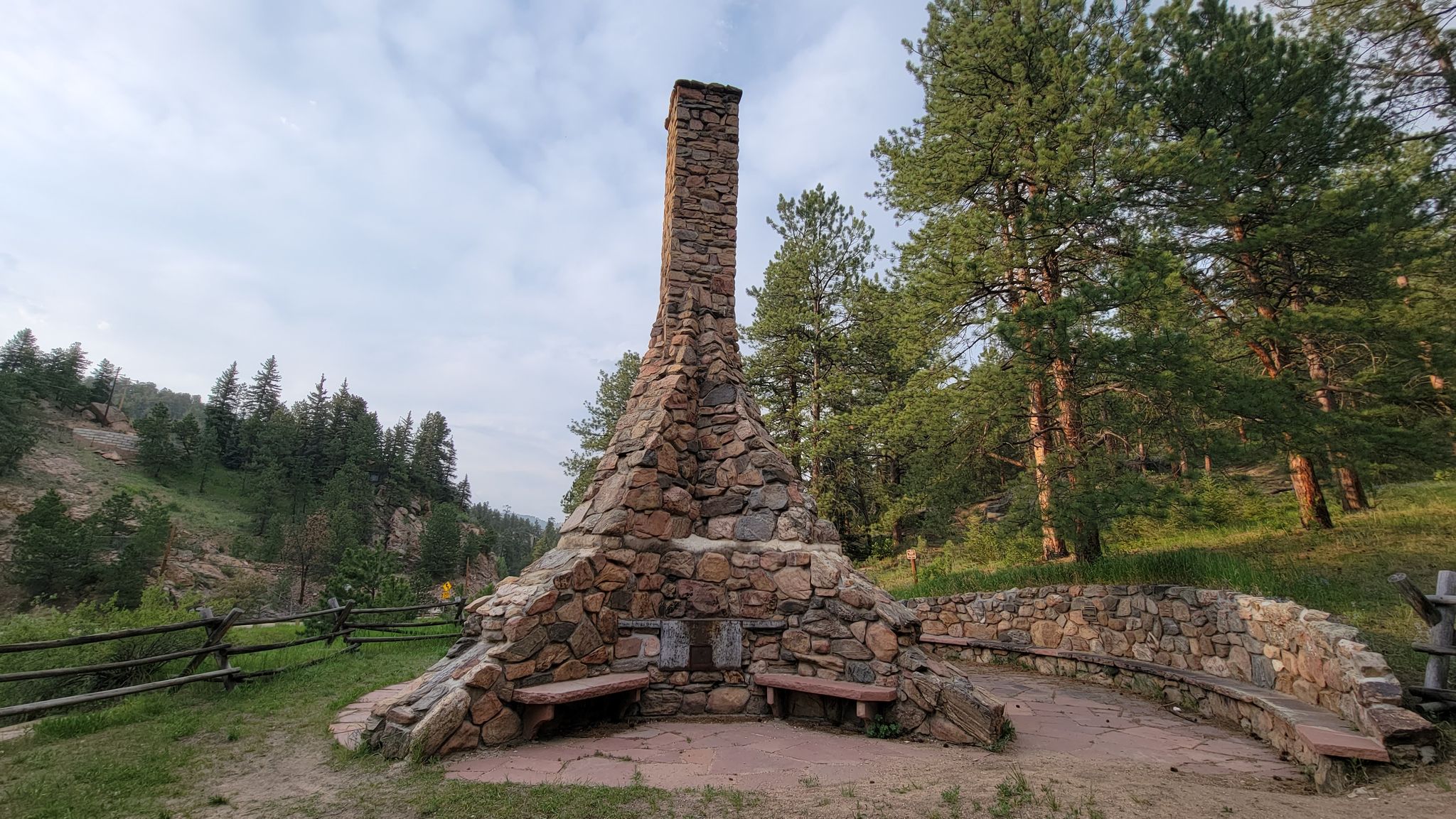 A tall stone chimney structure with benches, surrounded by pine trees. - Meadow View Loop