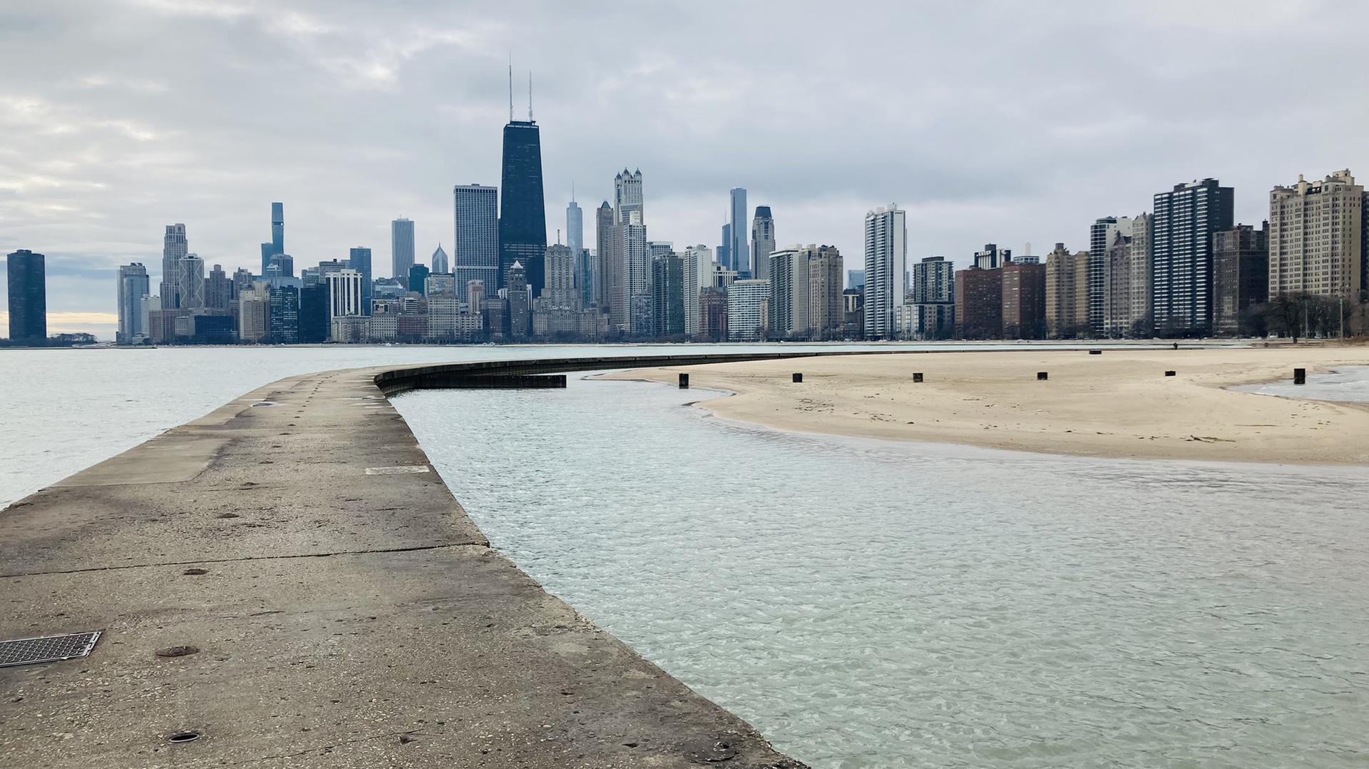 Concrete pier curves towards a city skyline, with beach and water under a cloudy sky.