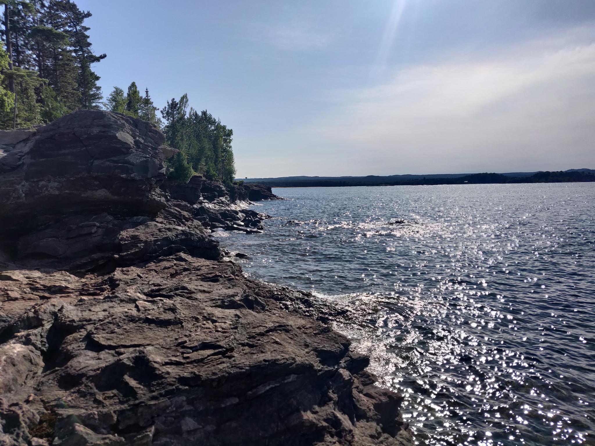 Sunlight sparkles on dark water next to a rugged, rocky shoreline and green trees.
