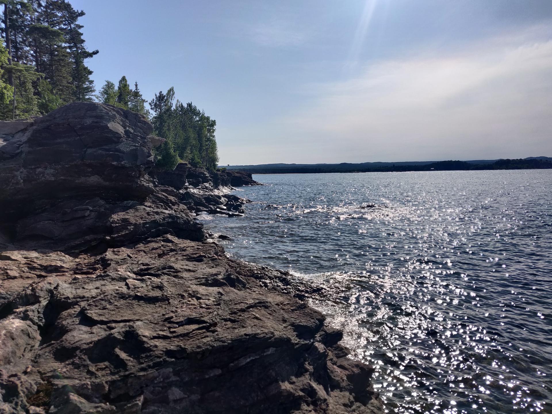Sunlight sparkles on dark water next to a rugged, rocky shoreline and green trees.
