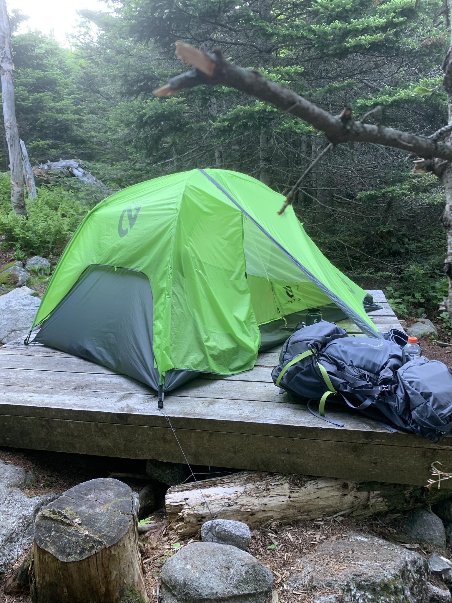 Bright green tent and backpack on a wooden platform in a shaded forest campsite.