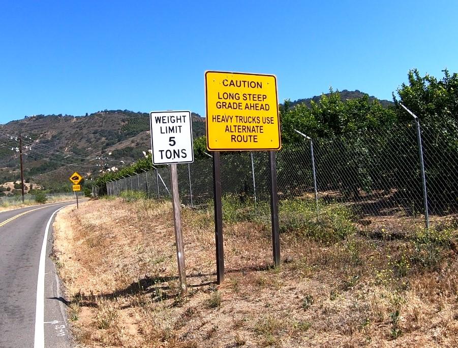 Two road signs next to an asphalt road with hills in background.
