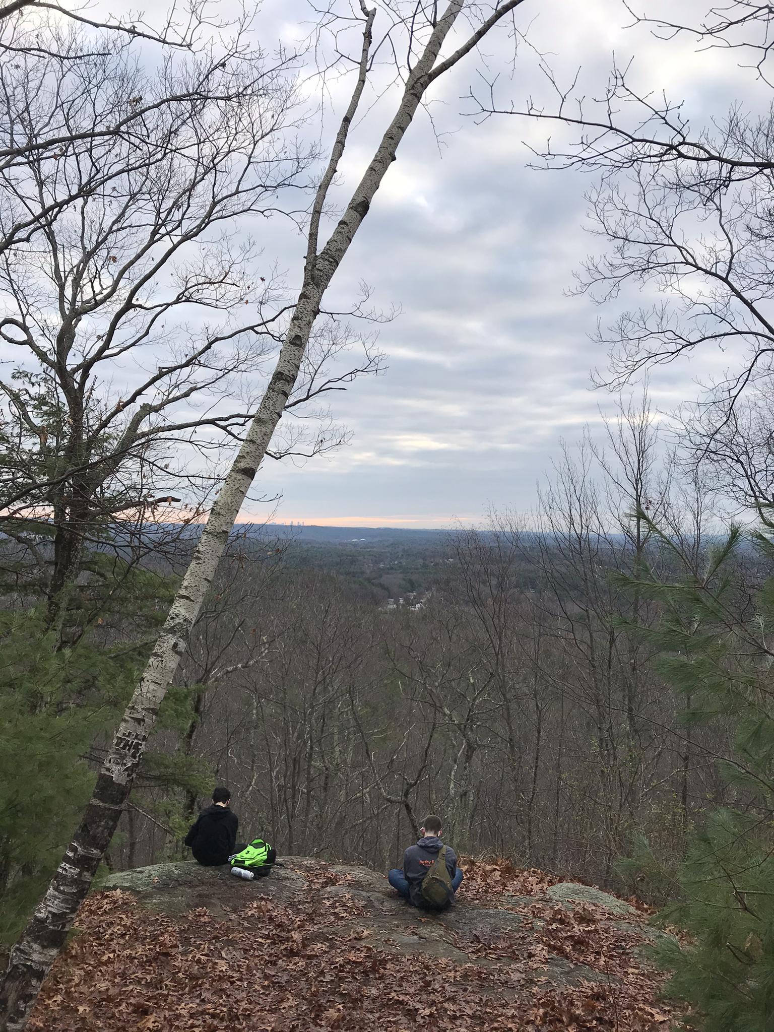Two people sit on a rocky ledge overlooking a vast winter forest.