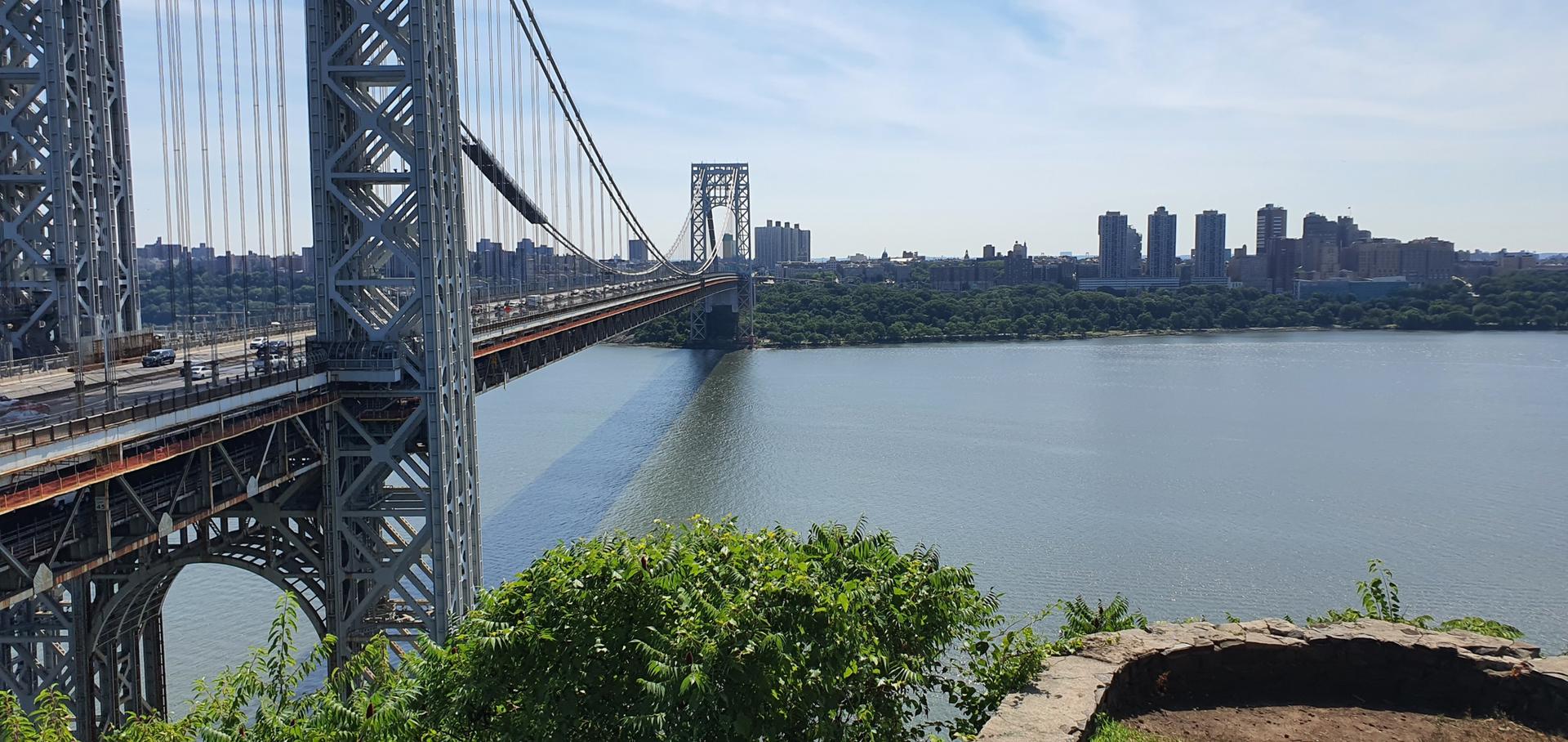 Suspension bridge over a wide river with a city skyline under bright sun.