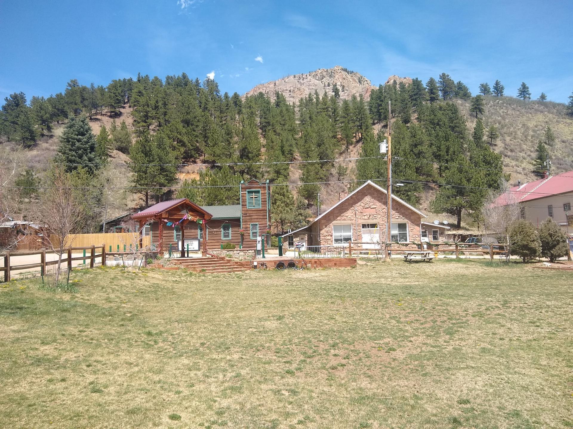 Small buildings in a sunny, grassy field with a pine-covered mountain backdrop.