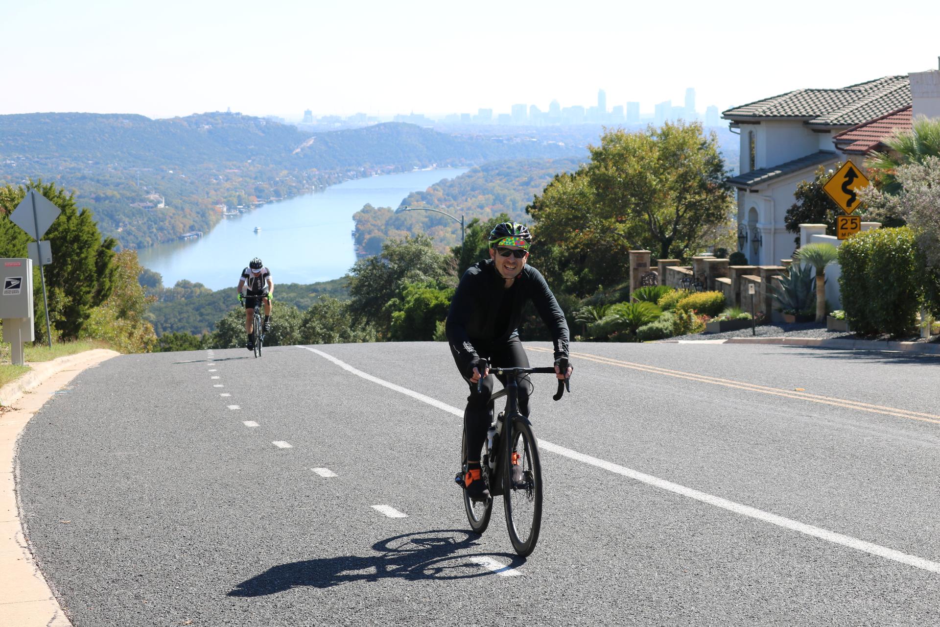 Two cyclists on a sunny, winding road overlooking a river and distant city.