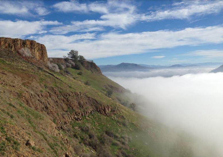 Sunny hillside and cliff face above a dense cloud inversion, distant mountains.