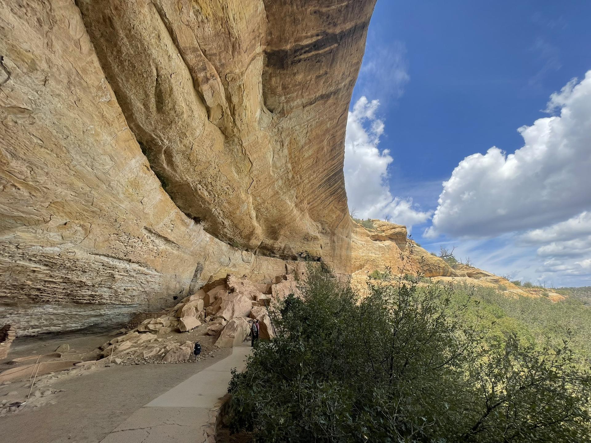 People walk on a path under a massive rock overhang, bright sky.