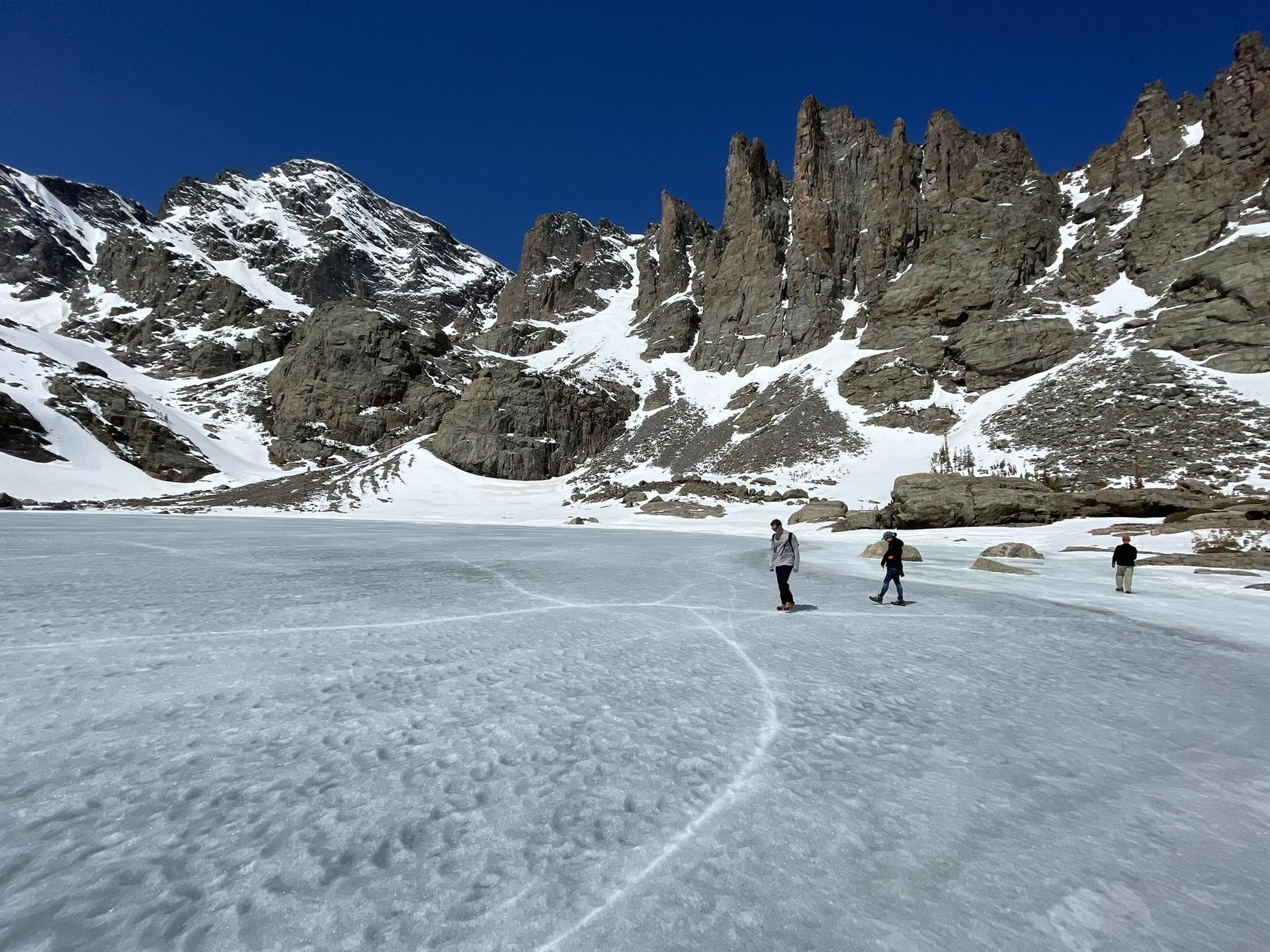 Three people walk across a vast frozen lake surrounded by snowy mountains.