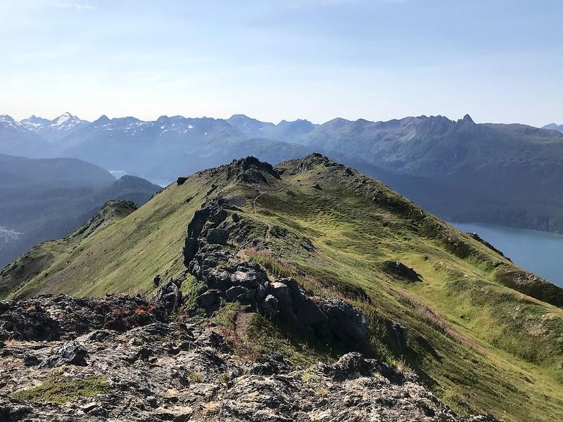 A rocky, grassy mountain ridge under a clear blue sky with distant peaks.