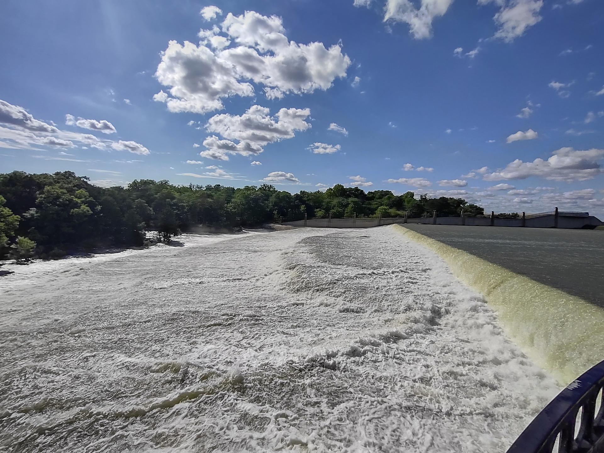 Rushing water flows over a wide dam with green trees under a blue sky.