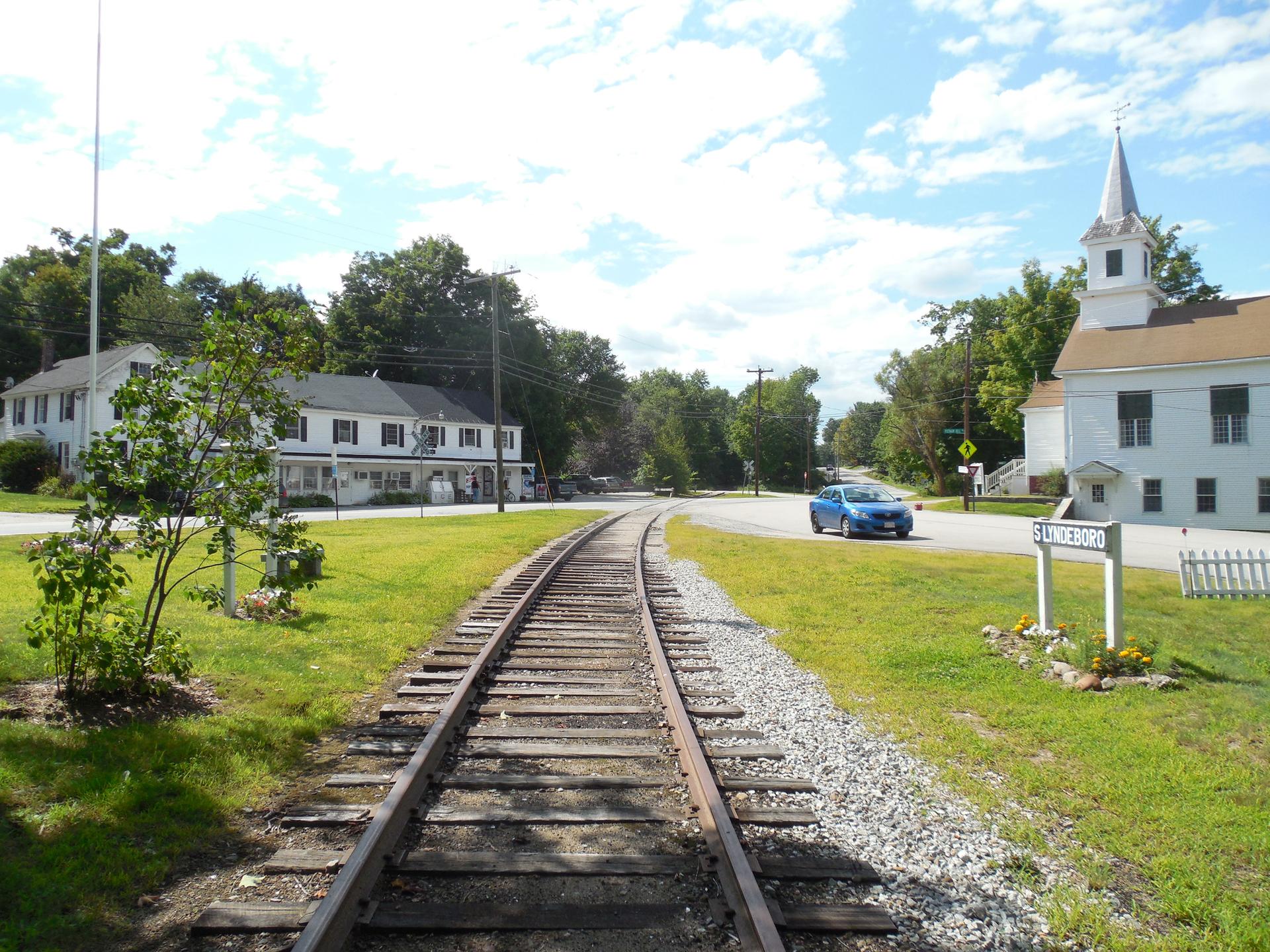 Sunny view of railroad tracks, buildings, a road with a car, and green grass.