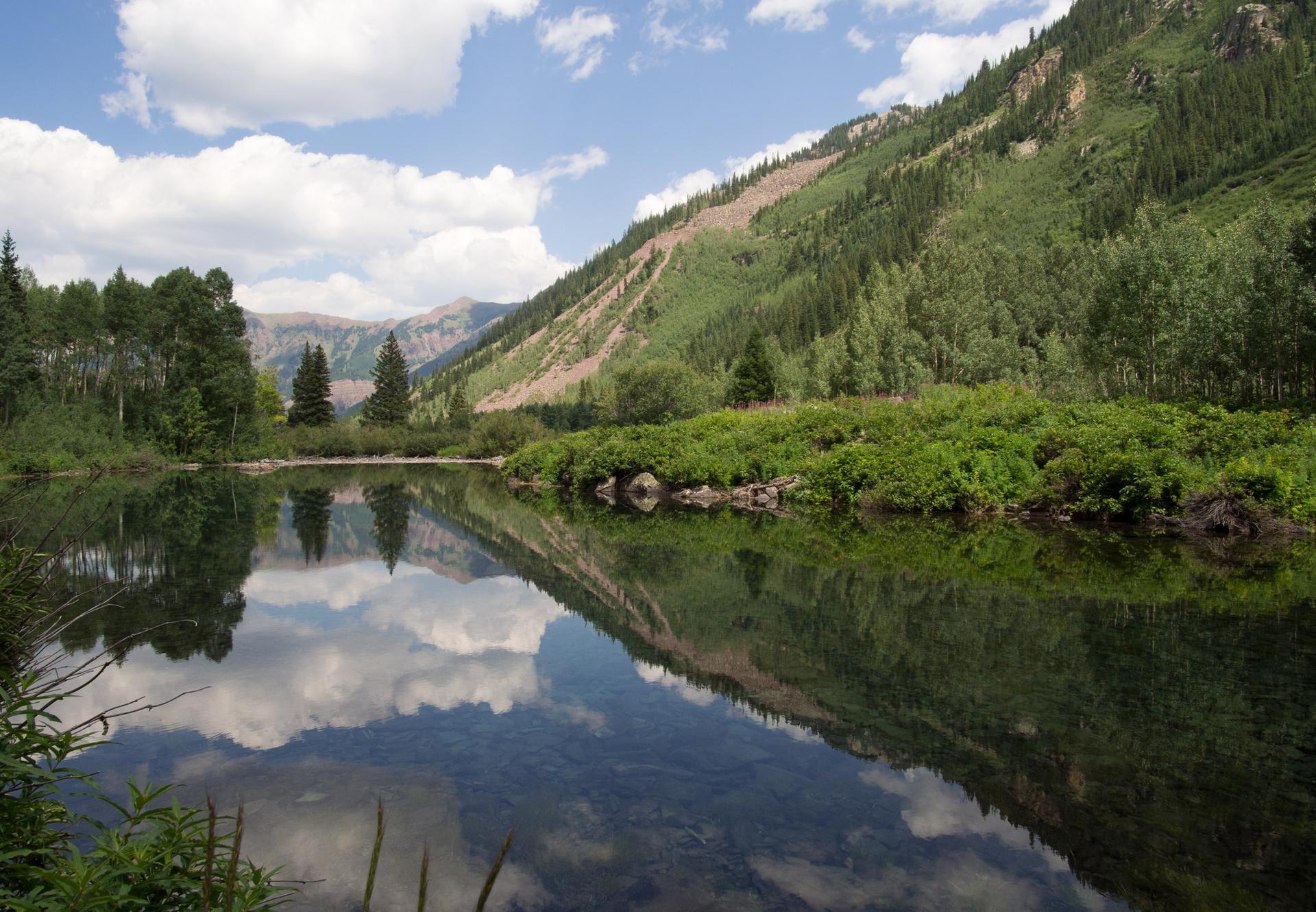 Calm lake reflecting forested mountains and cloudy blue sky on a bright day.