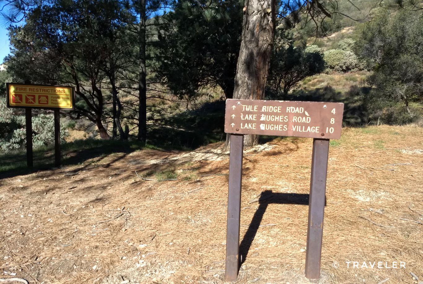 Two wooden trail signs stand on dry ground under trees on a sunny day.