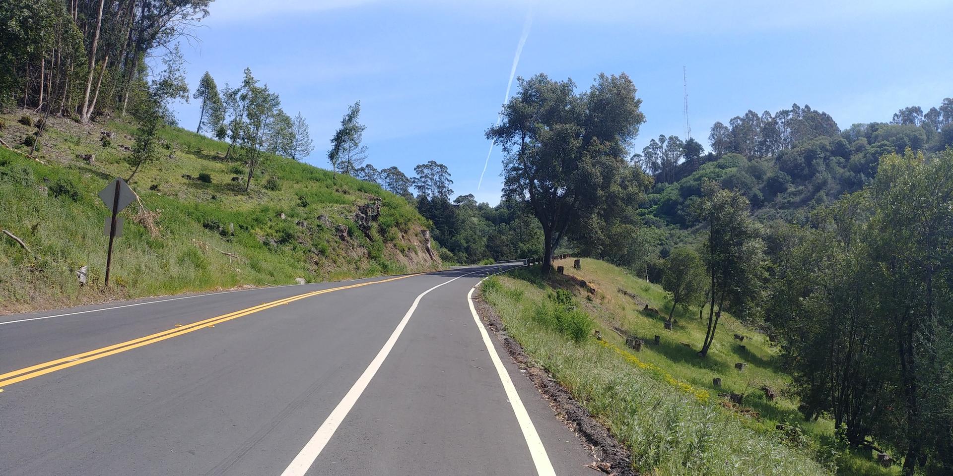 Winding road with a bike lane through green, tree-covered hills.