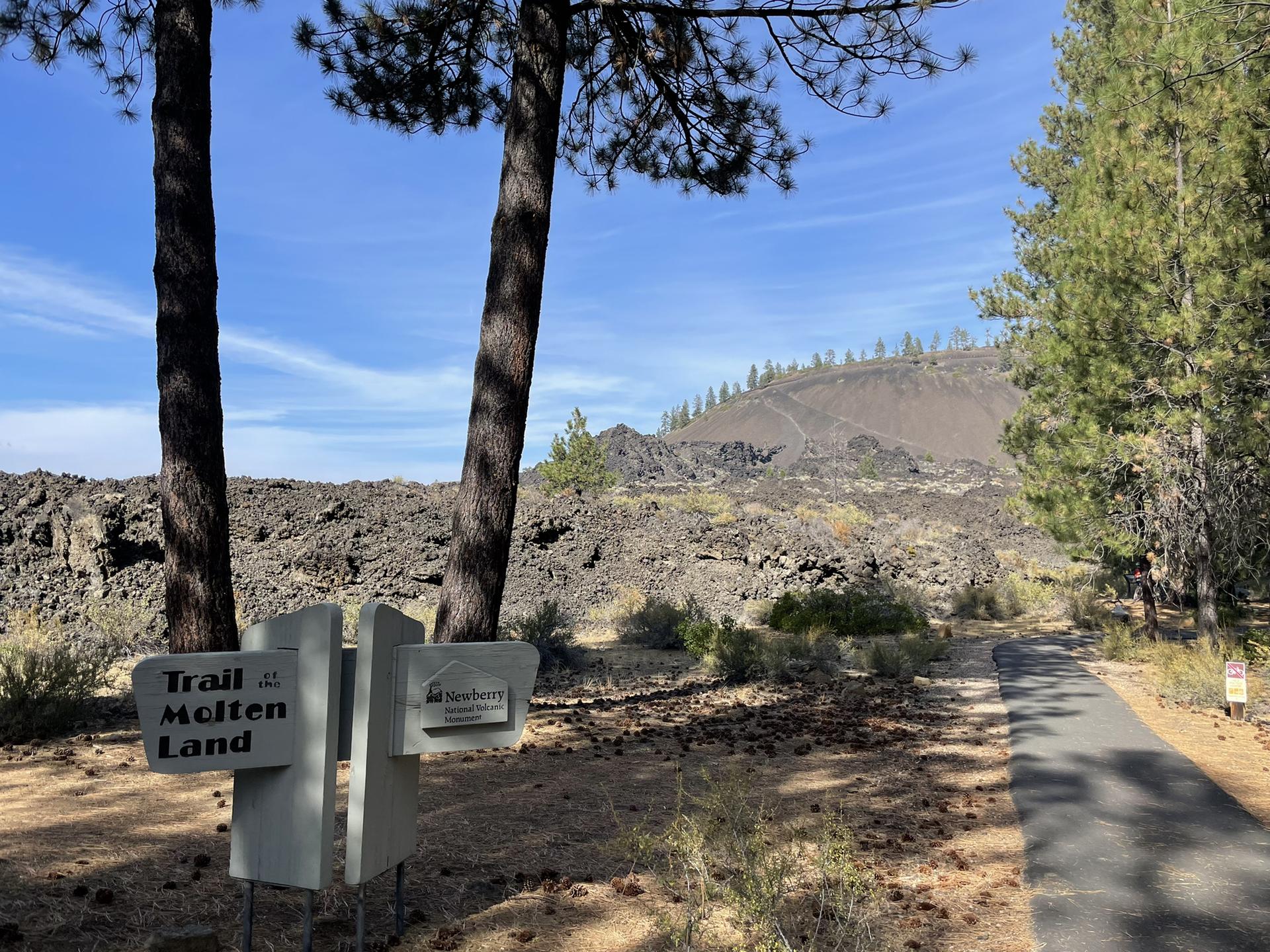 Trail signs next to a paved path, leading into a rocky, volcanic landscape.