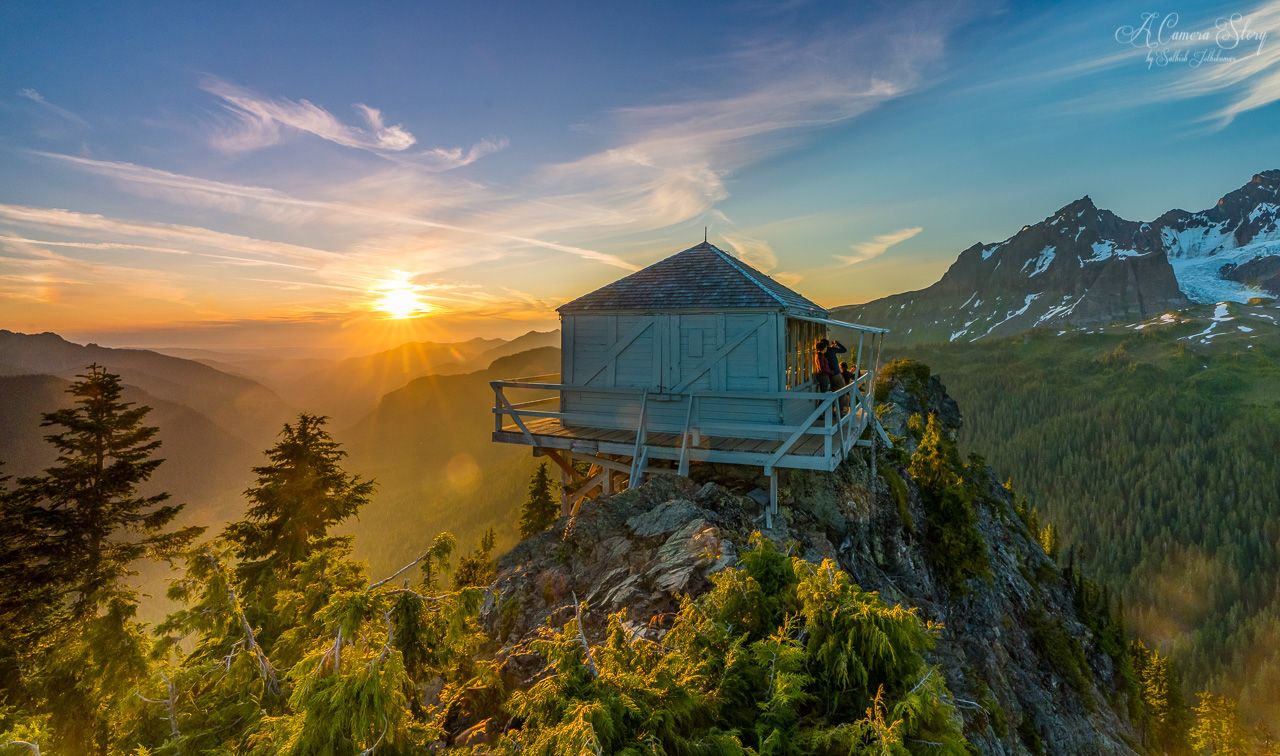 Lookout cabin on a mountain peak at sunset, with two people enjoying the view. - Park Butte Trail