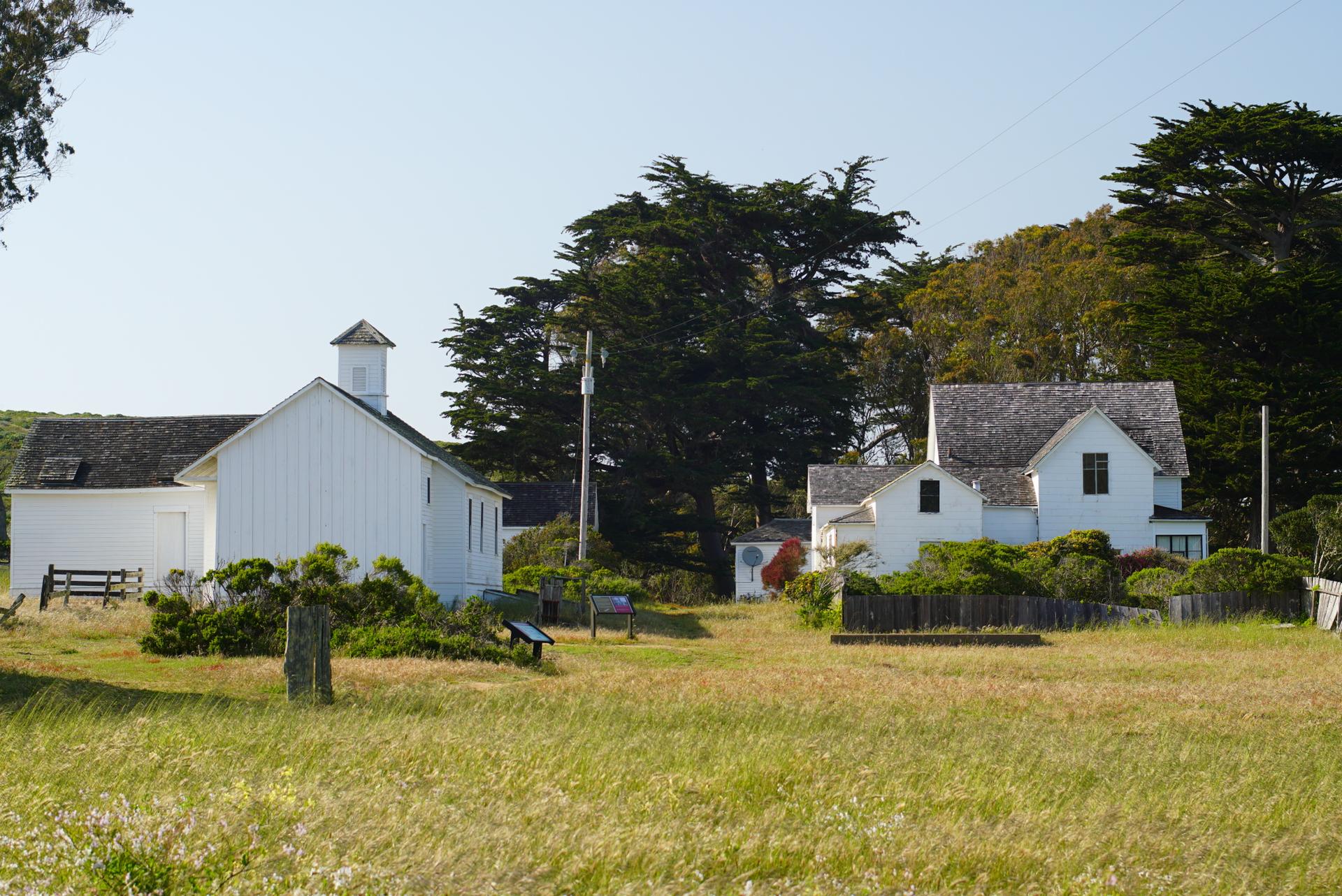 White historic buildings stand in a golden grassy field, framed by tall green trees.