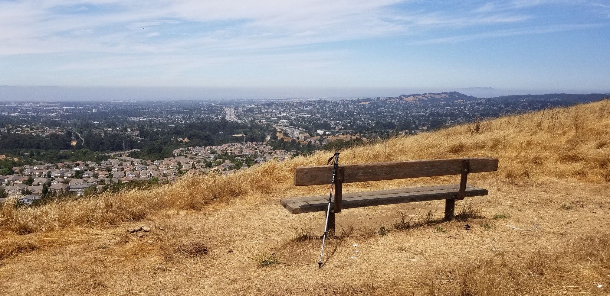 Wooden bench and trekking poles on a dry hillside overlooking a distant city. - Chabot-to-Garin Regional Trail