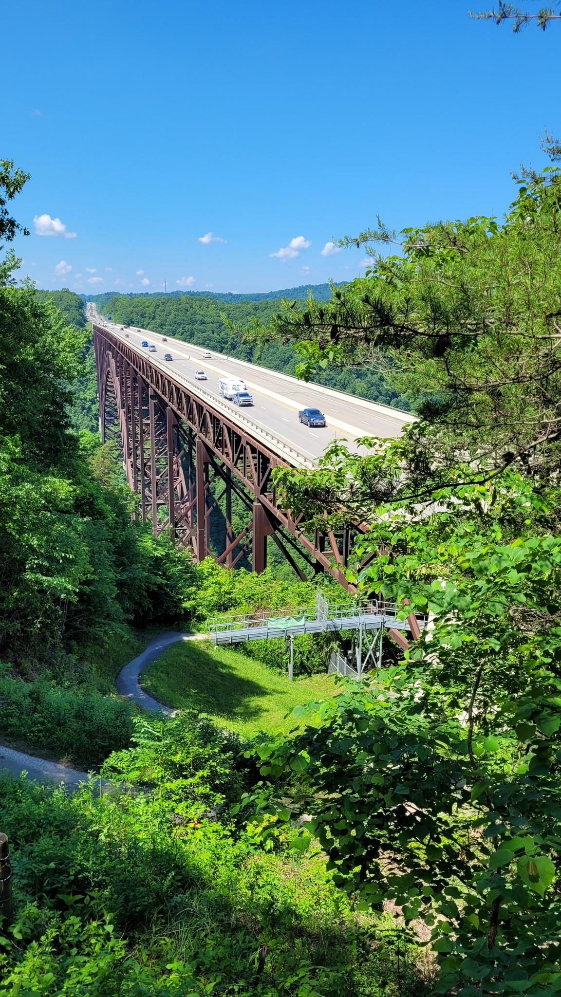 A long, tall bridge with cars crosses a green valley under a bright blue sky.