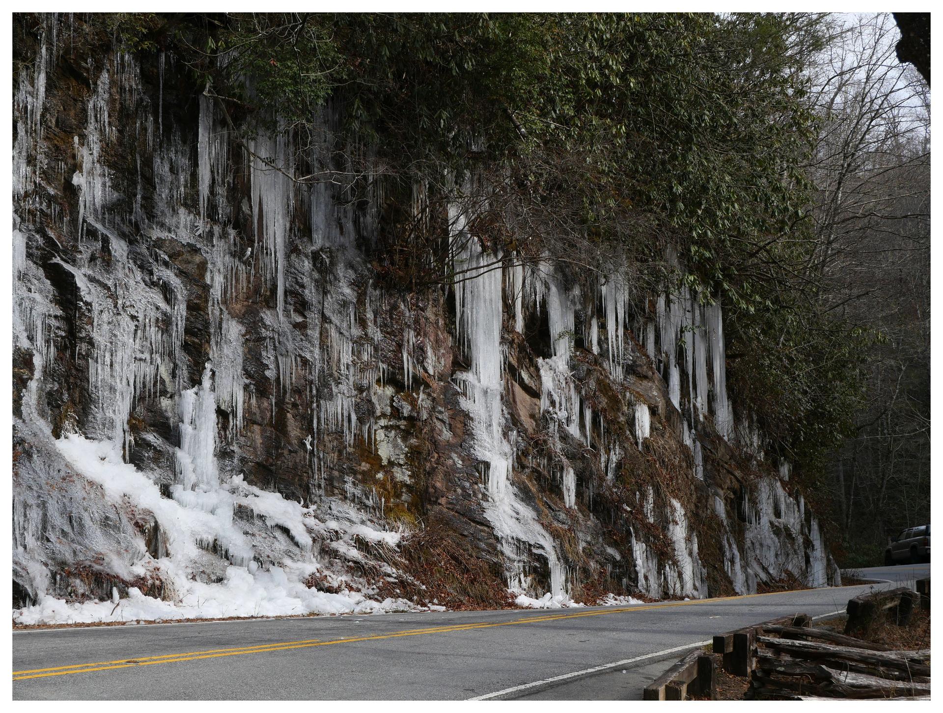 A rocky cliff face covered in long icicles and snow next to a road.