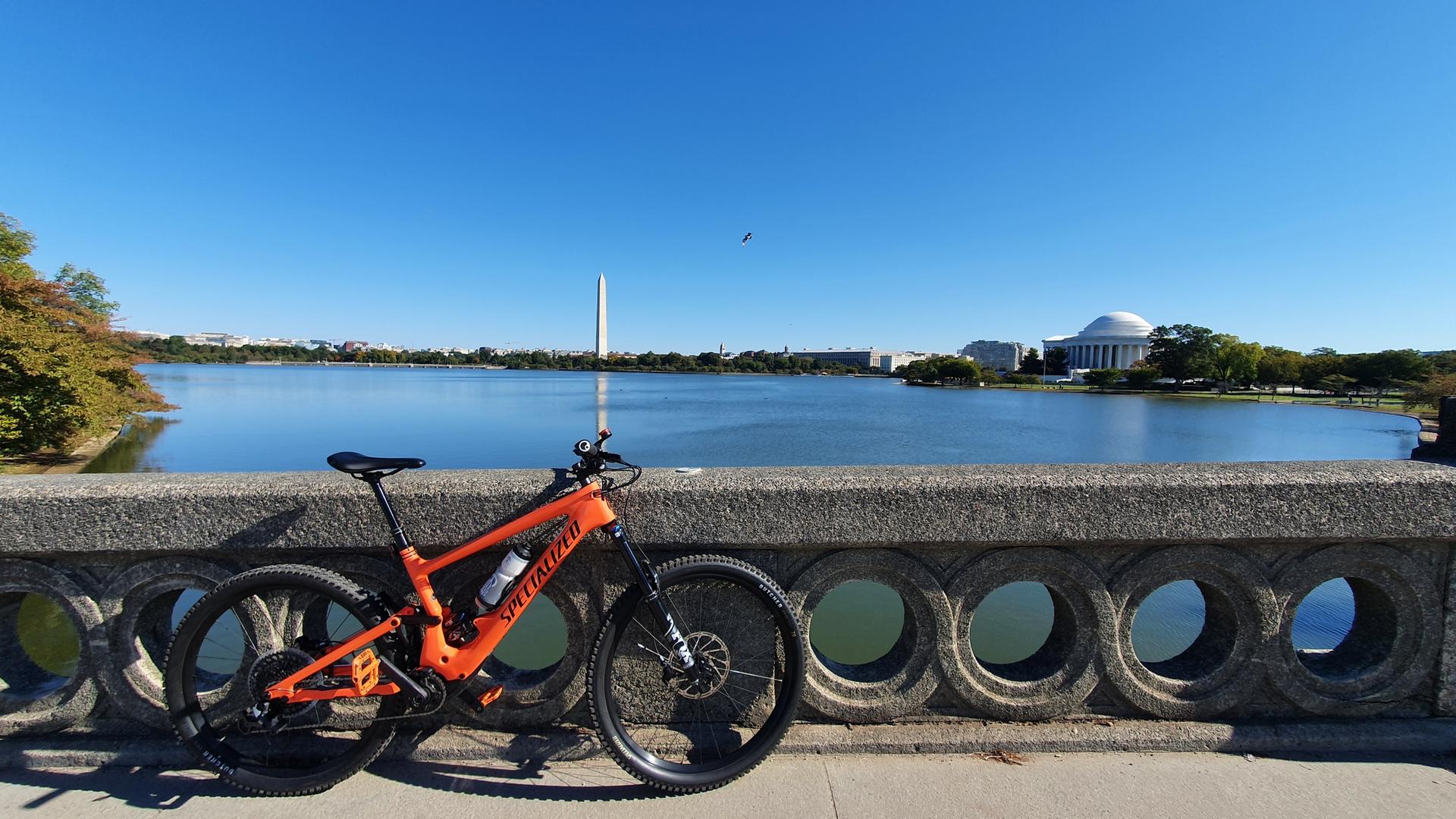 An orange mountain bike rests on a stone railing overlooking a lake and monuments.