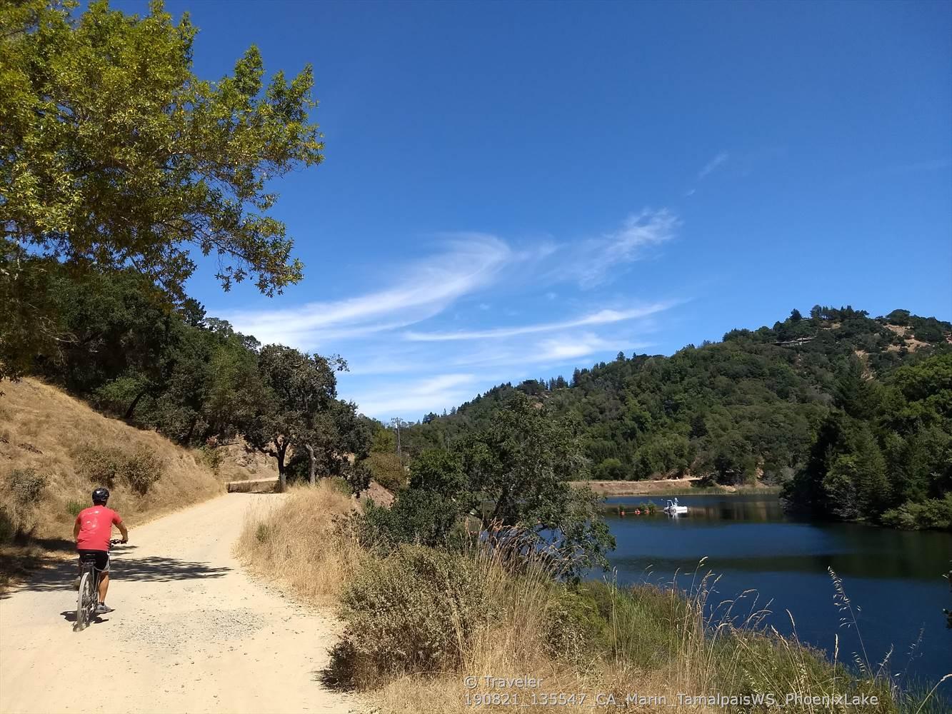 A person on a bike rides along a sunny dirt path beside a lake.