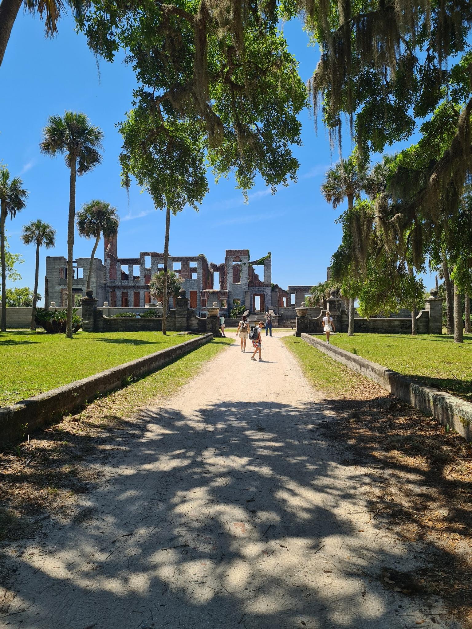 People walk on a sunny path towards large stone ruins surrounded by trees.