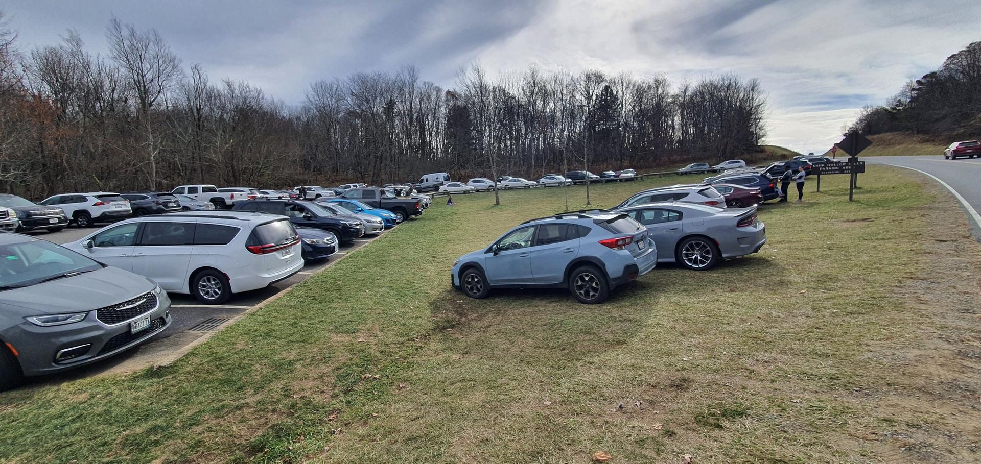 Crowded parking lot with many cars on a grassy hill under cloudy sky.