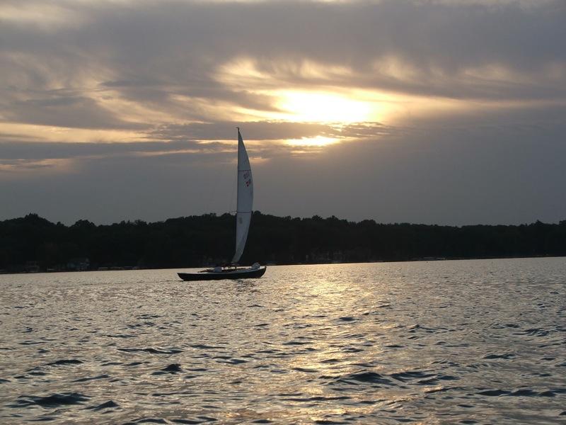 Sailboat silhouetted on water at sunset with cloudy sky and distant trees.