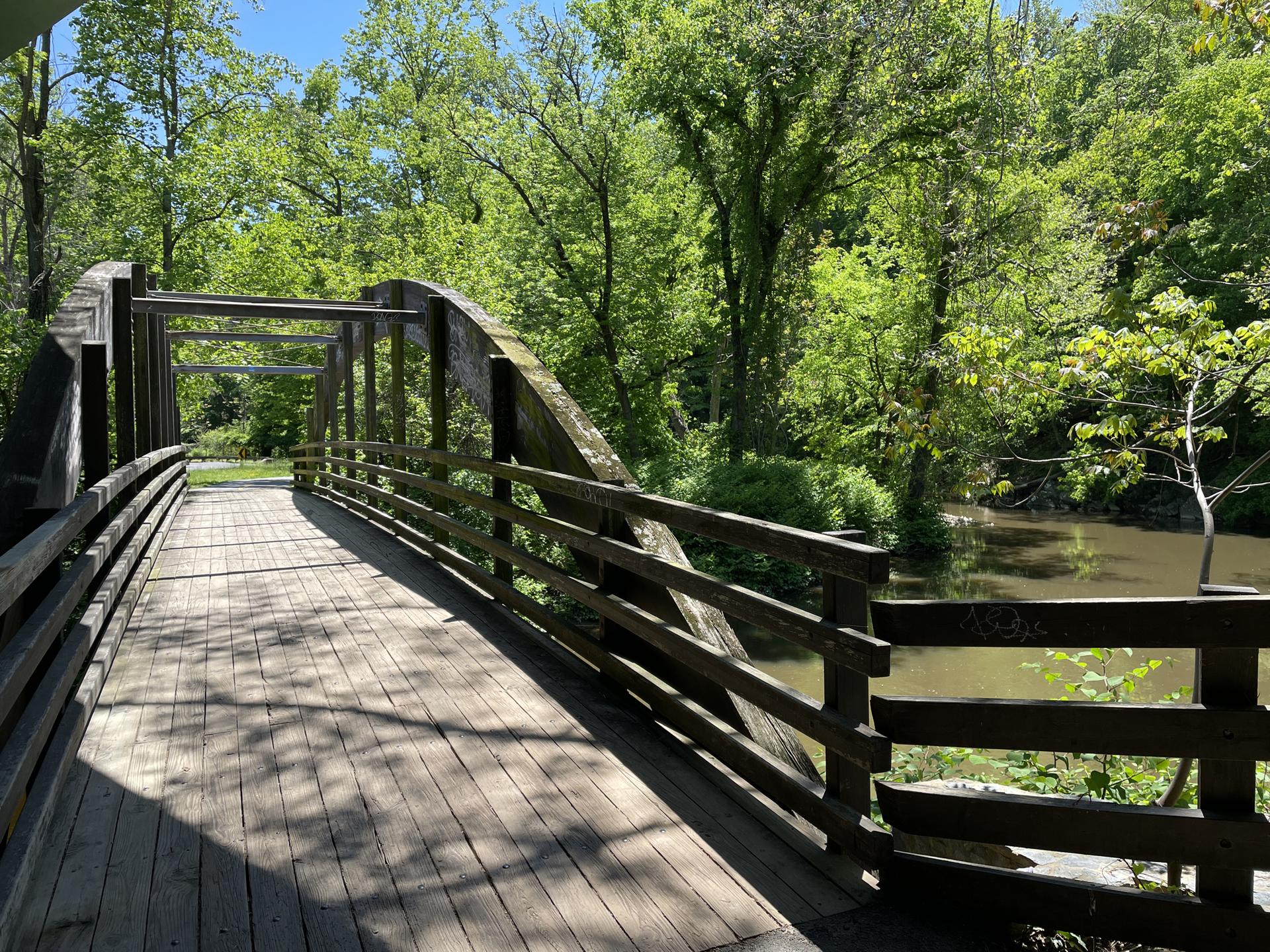 A wooden bridge spans a calm river, surrounded by lush green trees.