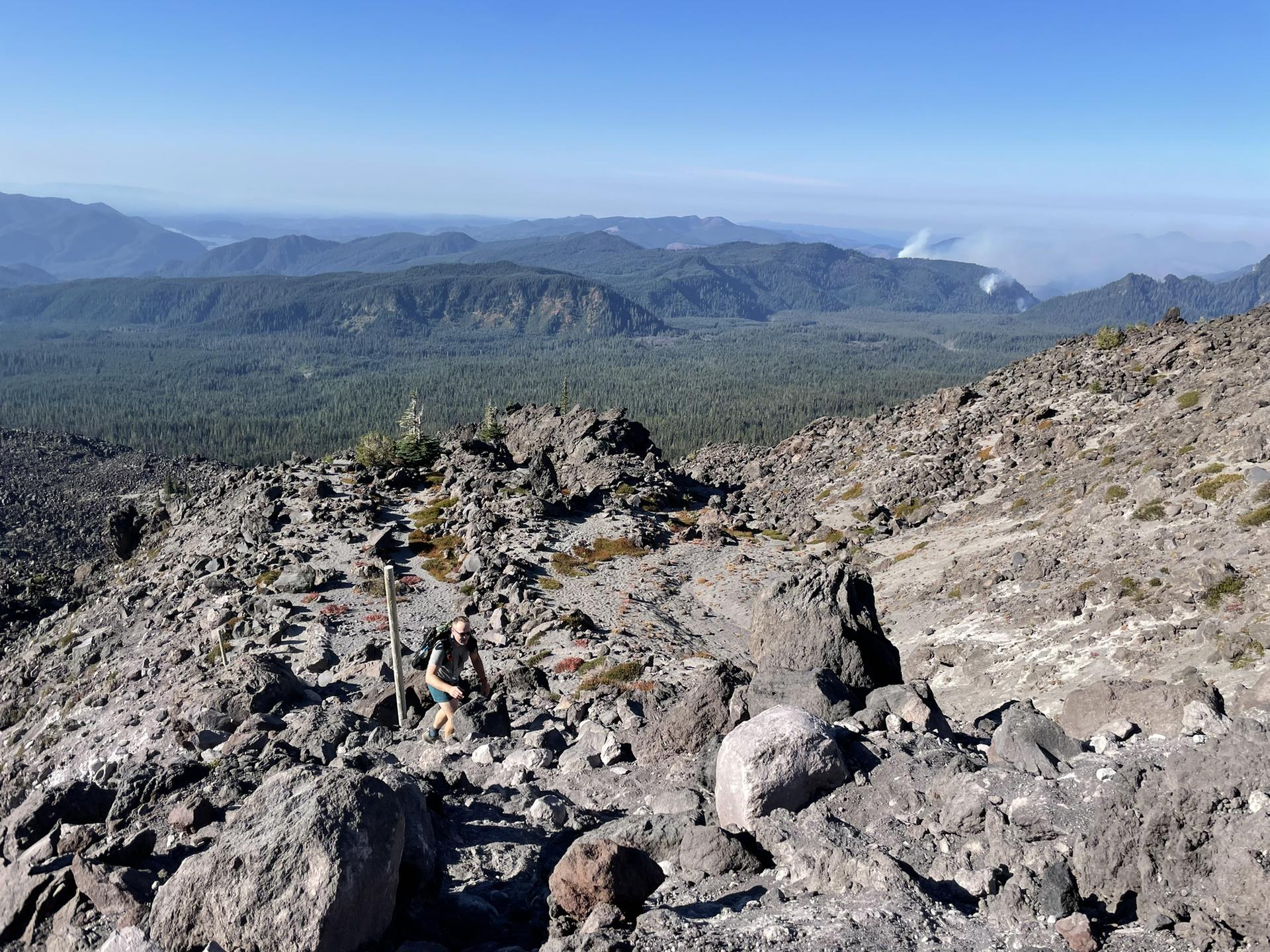 Hiker with poles ascending a rocky, barren mountain trail on a clear day.
