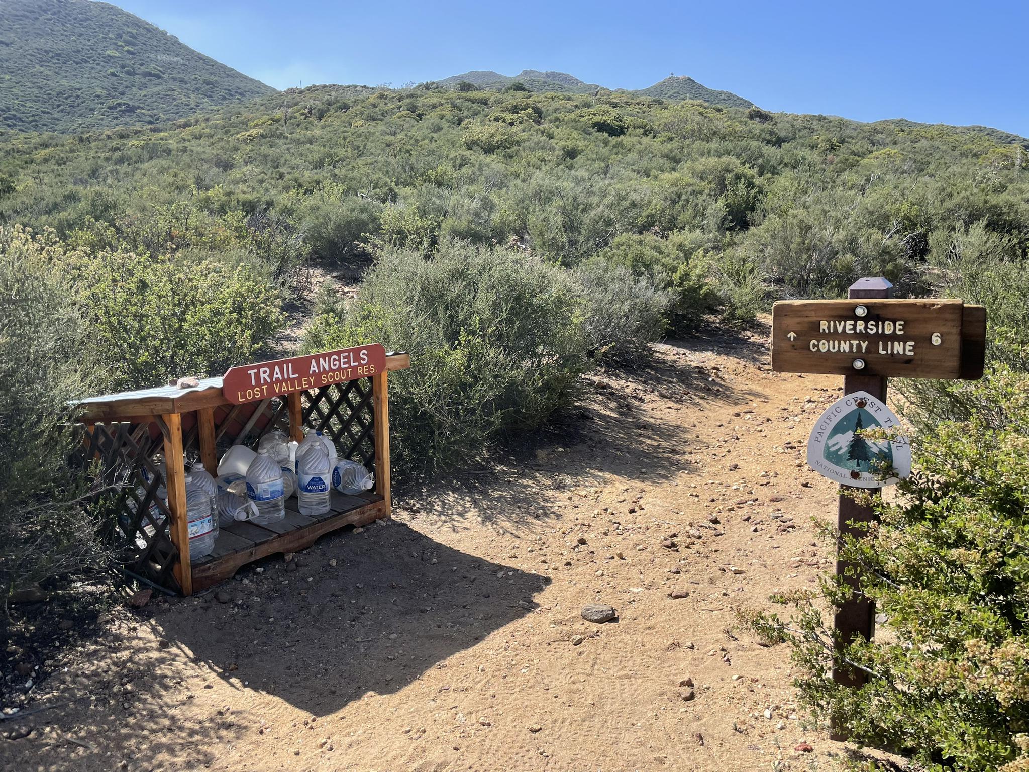 A trail sign and a wooden shelter with water bottles on a dirt path.