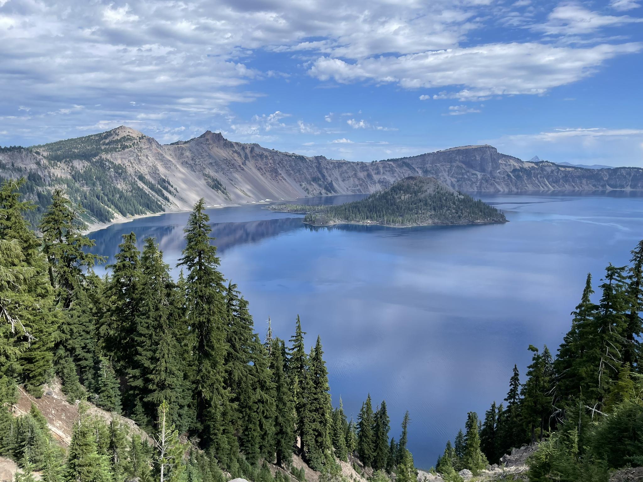 Expansive blue lake with a forested island, surrounded by mountains under a partly cloudy sky.