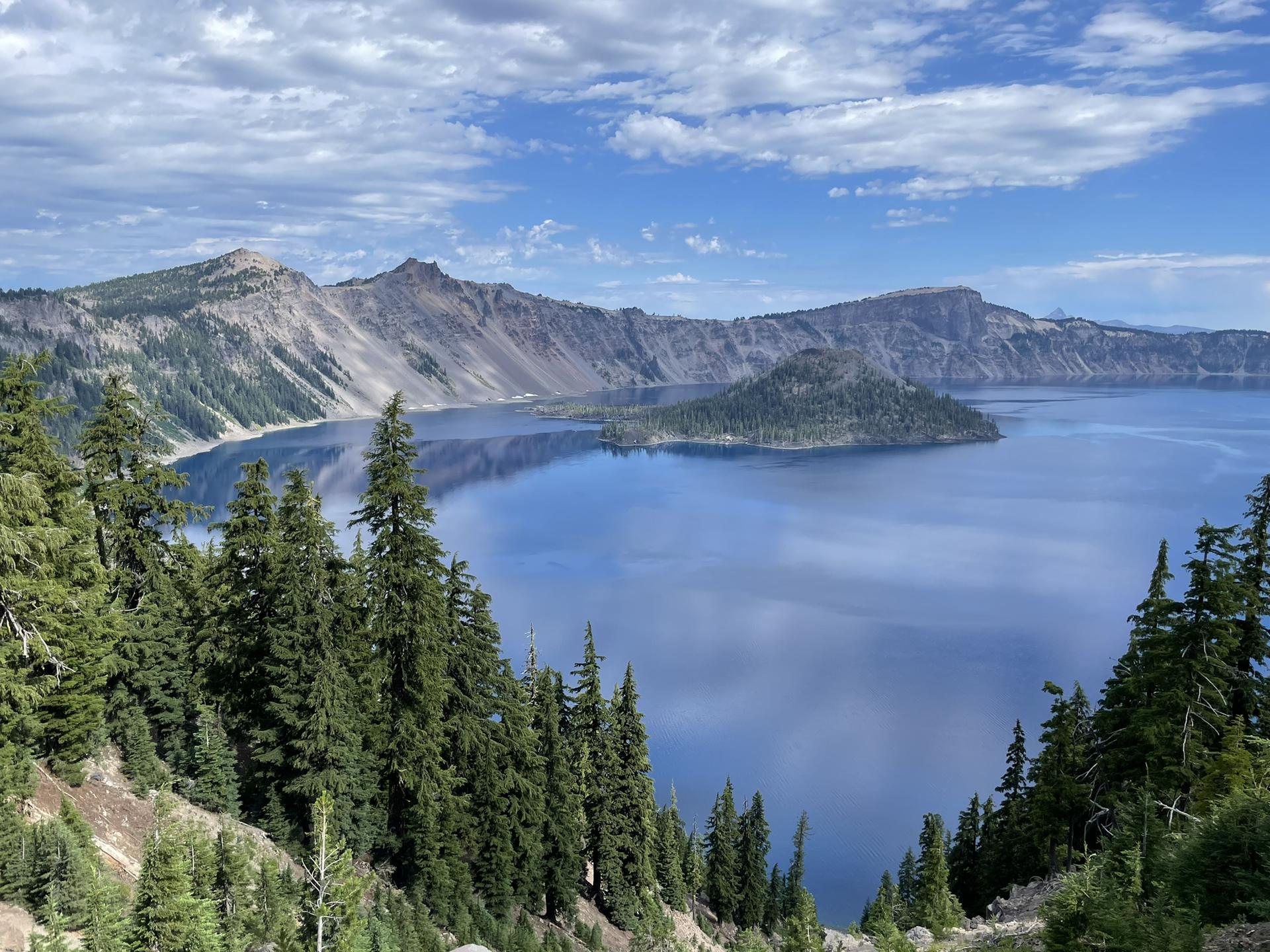 Expansive blue lake with a forested island, surrounded by mountains under a partly cloudy sky.