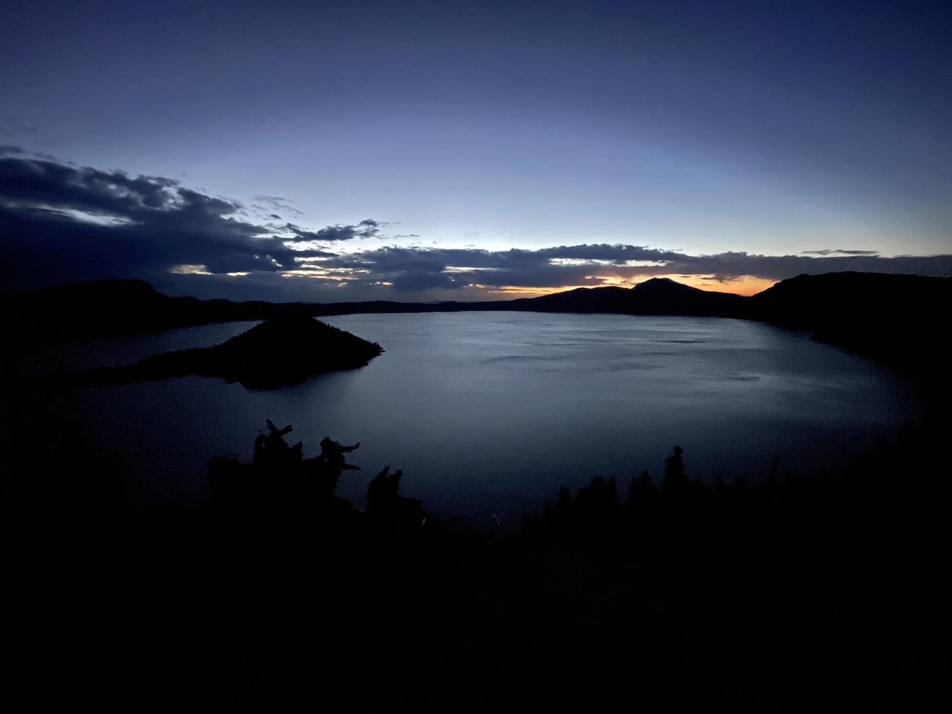 Dark twilight over a lake with an island, silhouetted mountains and cloudy sky.